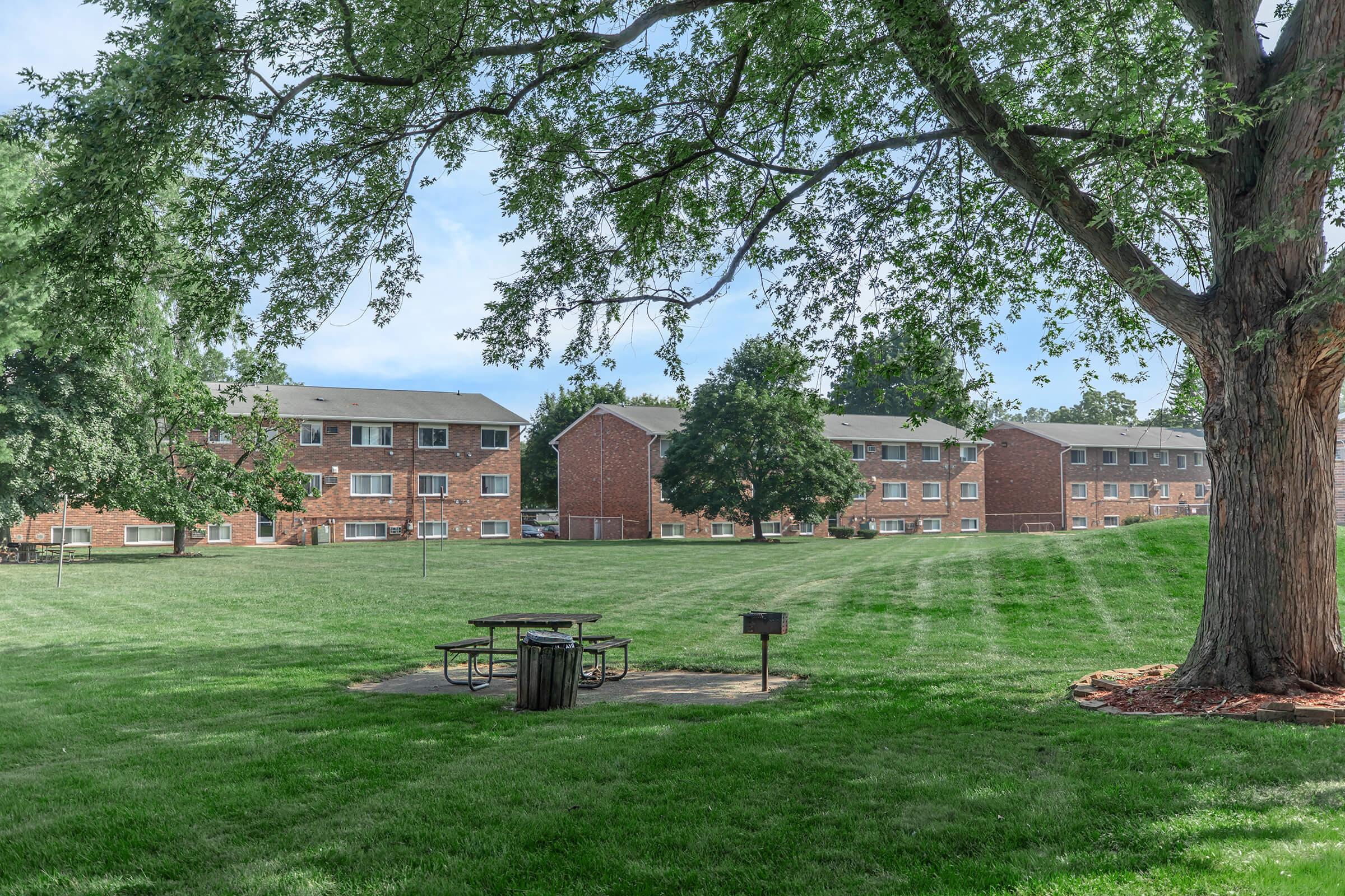 A grassy area with a large tree in the foreground, featuring a picnic table and a trash can. In the background, several brick apartment buildings are visible, surrounded by a well-maintained lawn under a clear sky.