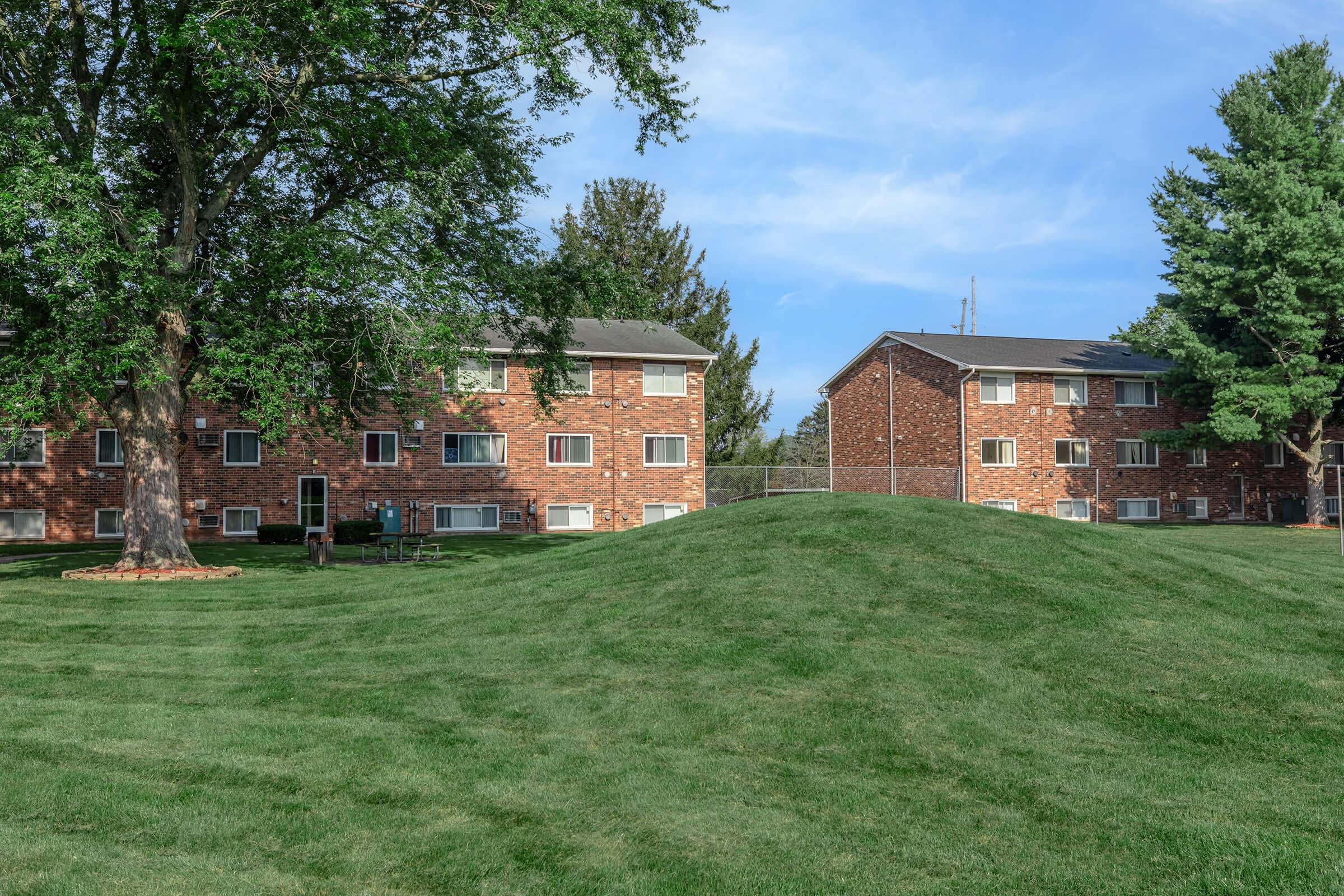 Two brick apartment buildings are situated on a grassy hill, surrounded by trees. The sky is clear with a few clouds. The grassy area features a neat lawn, and there's a sense of tranquility in the residential setting.