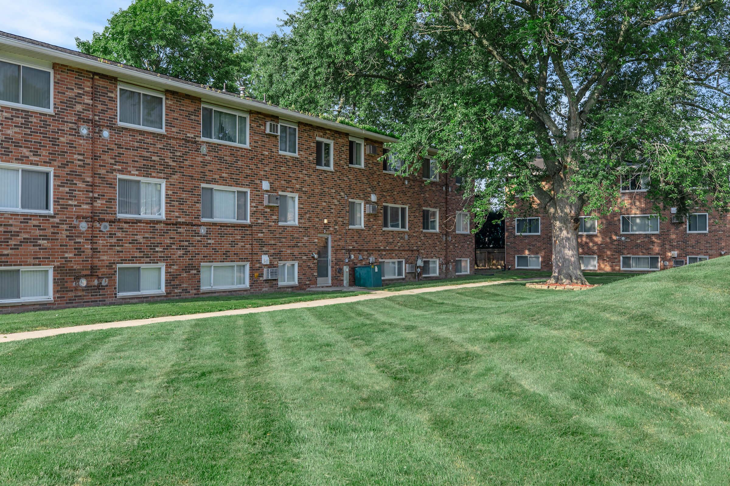 A brick apartment building with several windows, surrounded by well-maintained green grass and trees. The pathway in front of the building leads to a grassy area, creating a pleasant outdoor space.