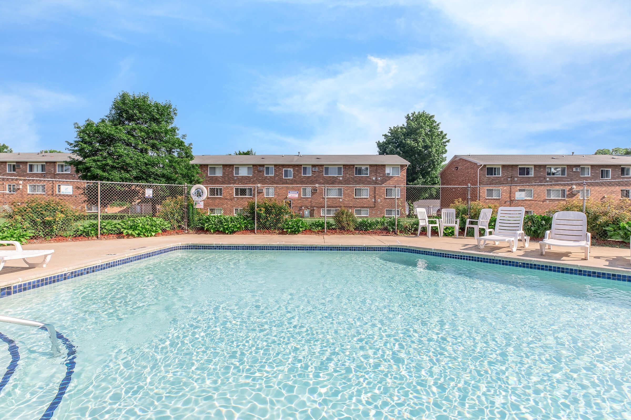 A clear swimming pool surrounded by a paved area with white lounge chairs, set against a backdrop of brick apartment buildings and lush greenery. The sky is bright blue with a few clouds, creating a sunny, inviting atmosphere.