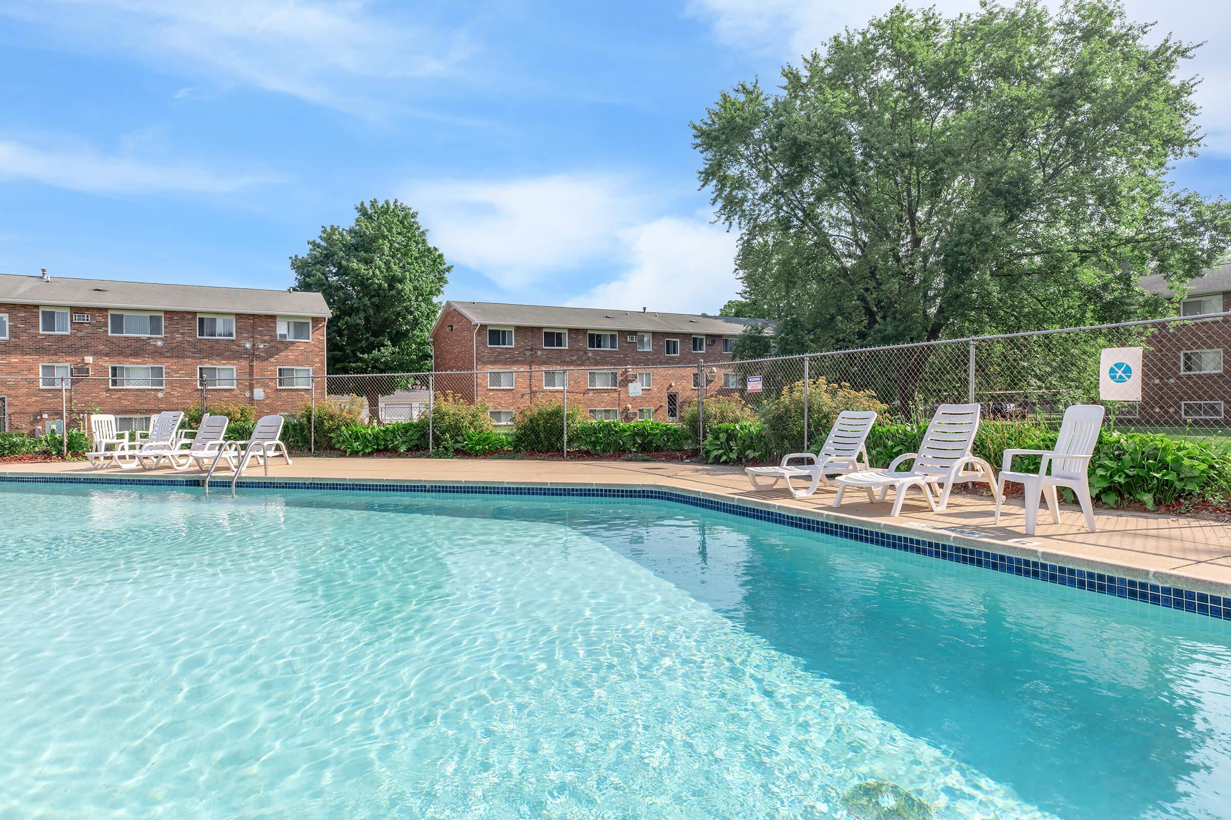 A clear blue pool surrounded by lounge chairs, with a lush green landscape and brick apartment buildings in the background. The scene is bright and inviting, showcasing a well-maintained outdoor area ideal for relaxation.