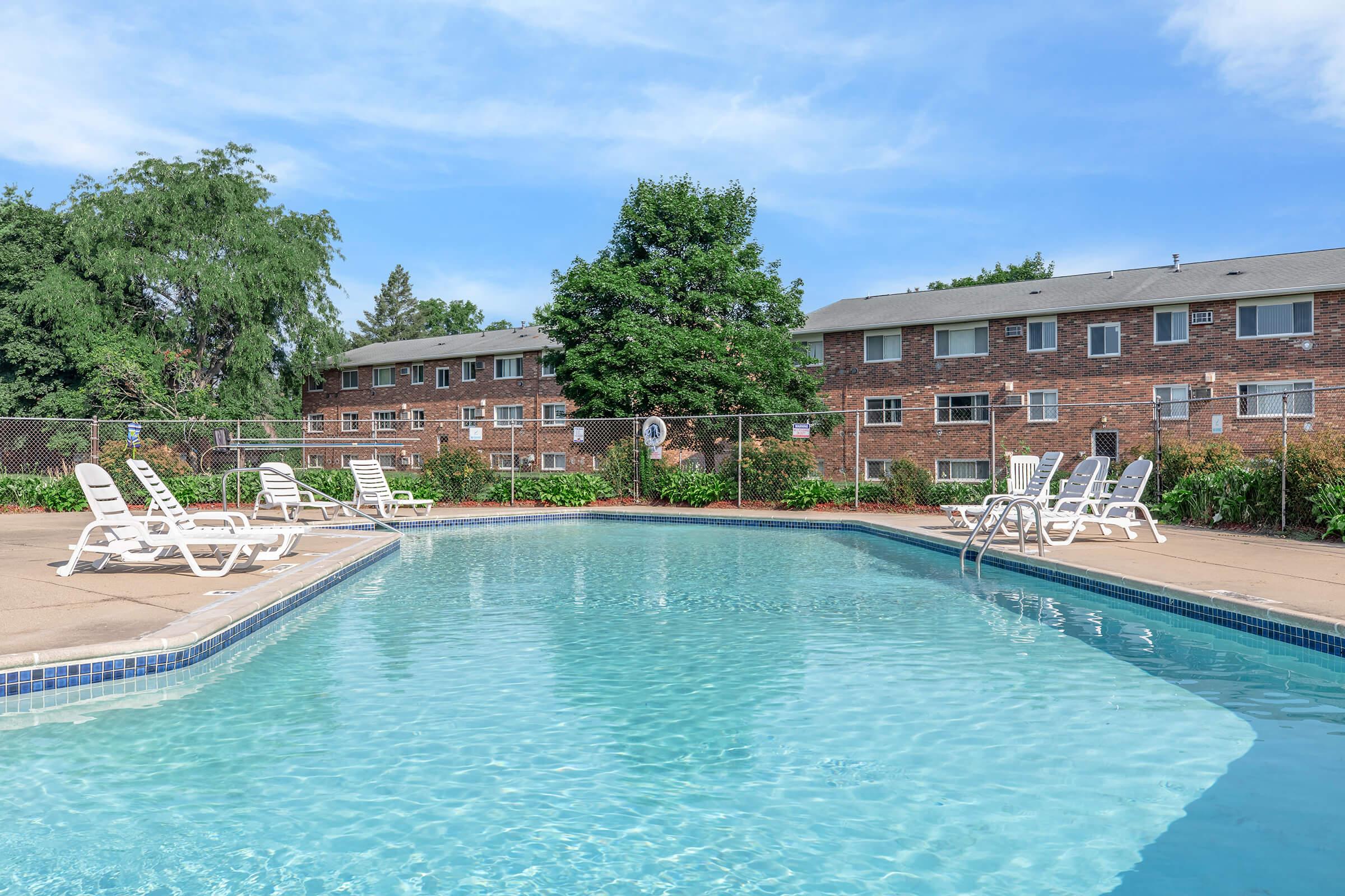 A clear swimming pool surrounded by lounge chairs, with a backdrop of brick apartment buildings and greenery. The sky is blue with a few clouds, creating a bright and inviting atmosphere. The area is well-maintained, perfect for relaxation and leisure.