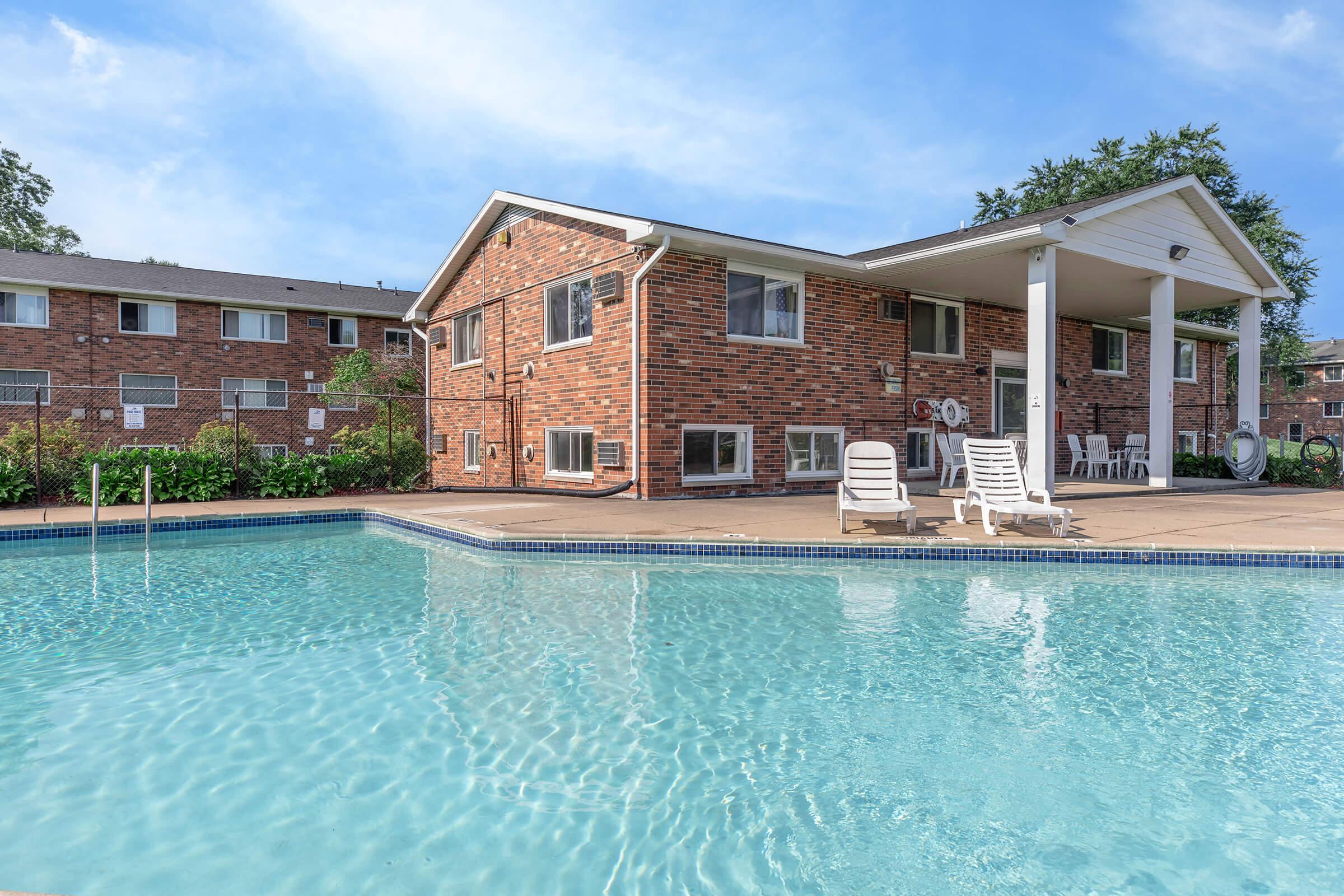 A clear swimming pool in front of a brick apartment building, with lounge chairs arranged around the pool area. Lush green plants and trees can be seen in the background, and a blue sky adds to the inviting atmosphere.