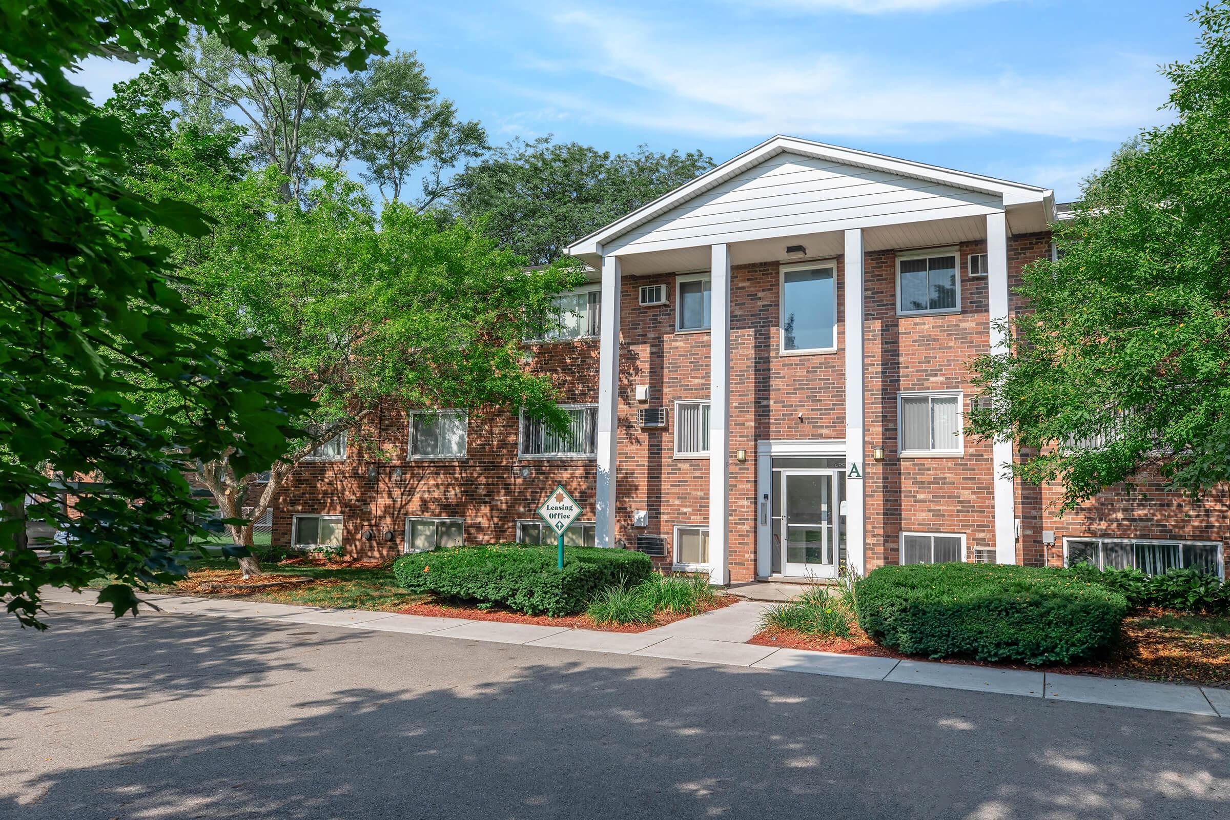 A three-story brick apartment building surrounded by green trees and shrubs. The entrance features a glass door flanked by large windows. A paved pathway leads to the entrance, and a sign is visible near the entrance. The sky is clear with some clouds.