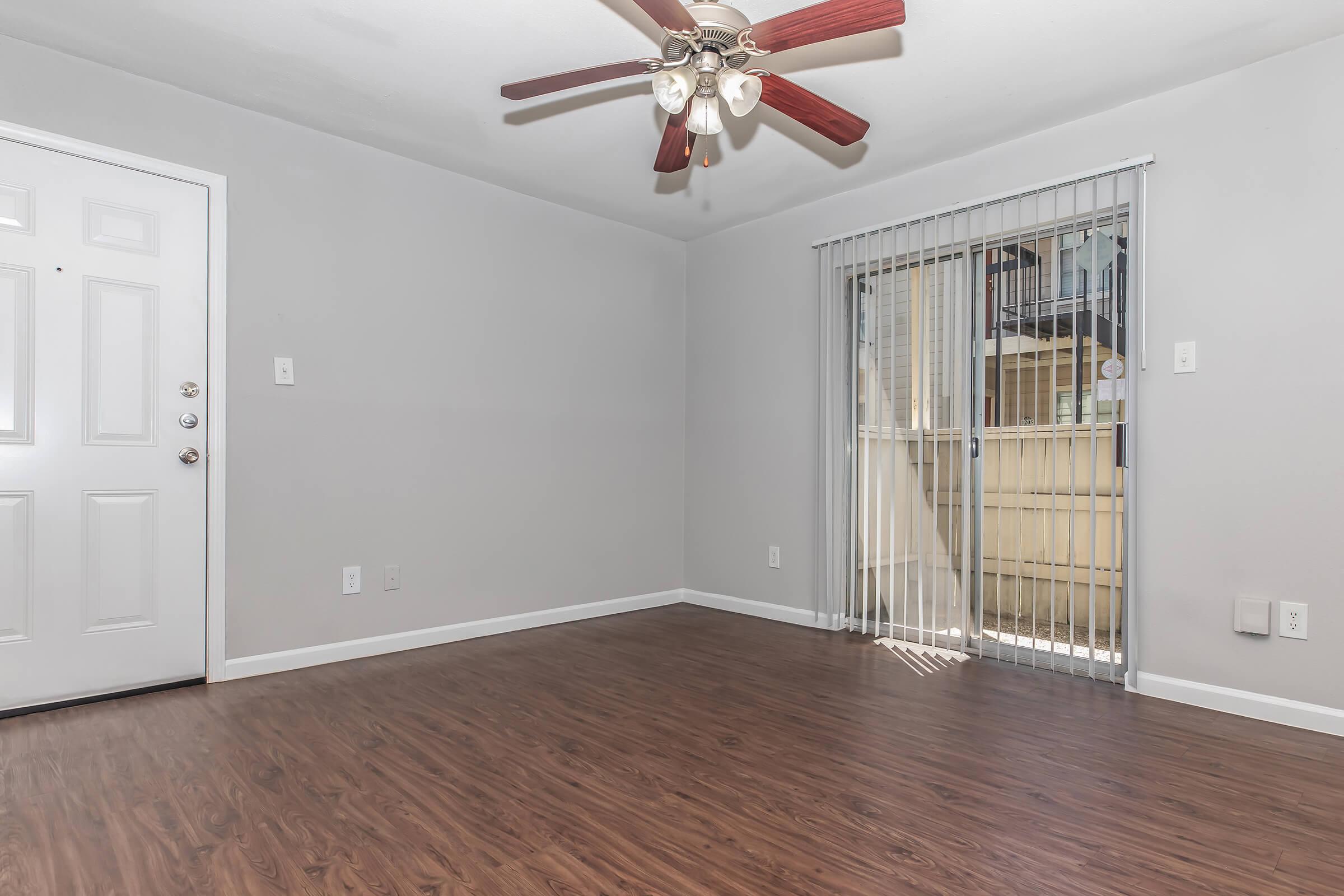 A well-lit, empty living room featuring light gray walls, wooden floors, and a ceiling fan with wooden blades. The room includes a front door on the left and sliding glass doors on the right, leading to a balcony. The space is clean and ready for furniture.