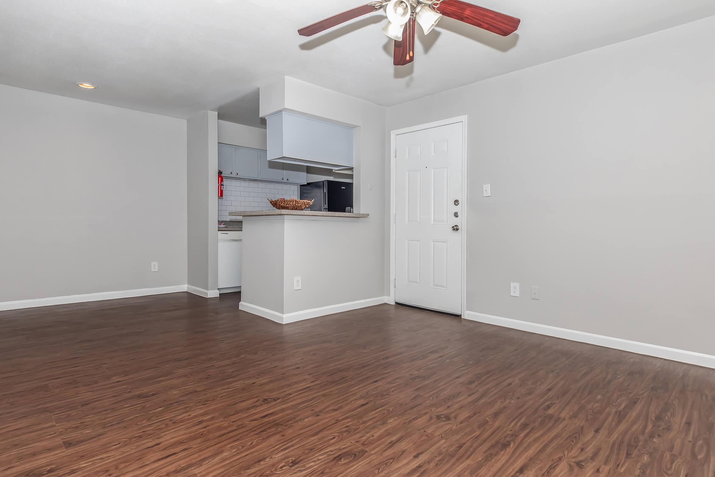 Spacious, open living area with light-colored walls and hardwood-style flooring. A ceiling fan is visible, and a doorway leads to the kitchen area featuring modern appliances. The entrance door is positioned on one wall, creating a welcoming atmosphere. Ideal for comfortable living.