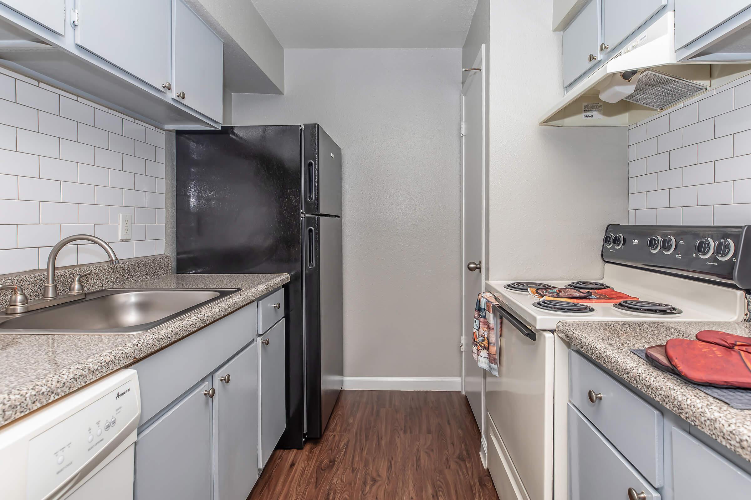 A modern kitchen featuring light blue cabinets, a black refrigerator, and a stainless steel sink. The countertop is made of granite, and there's a white tiled backsplash. The kitchen also includes a stove with an oven and red oven mitts hanging nearby, all set against a neutral wall color.