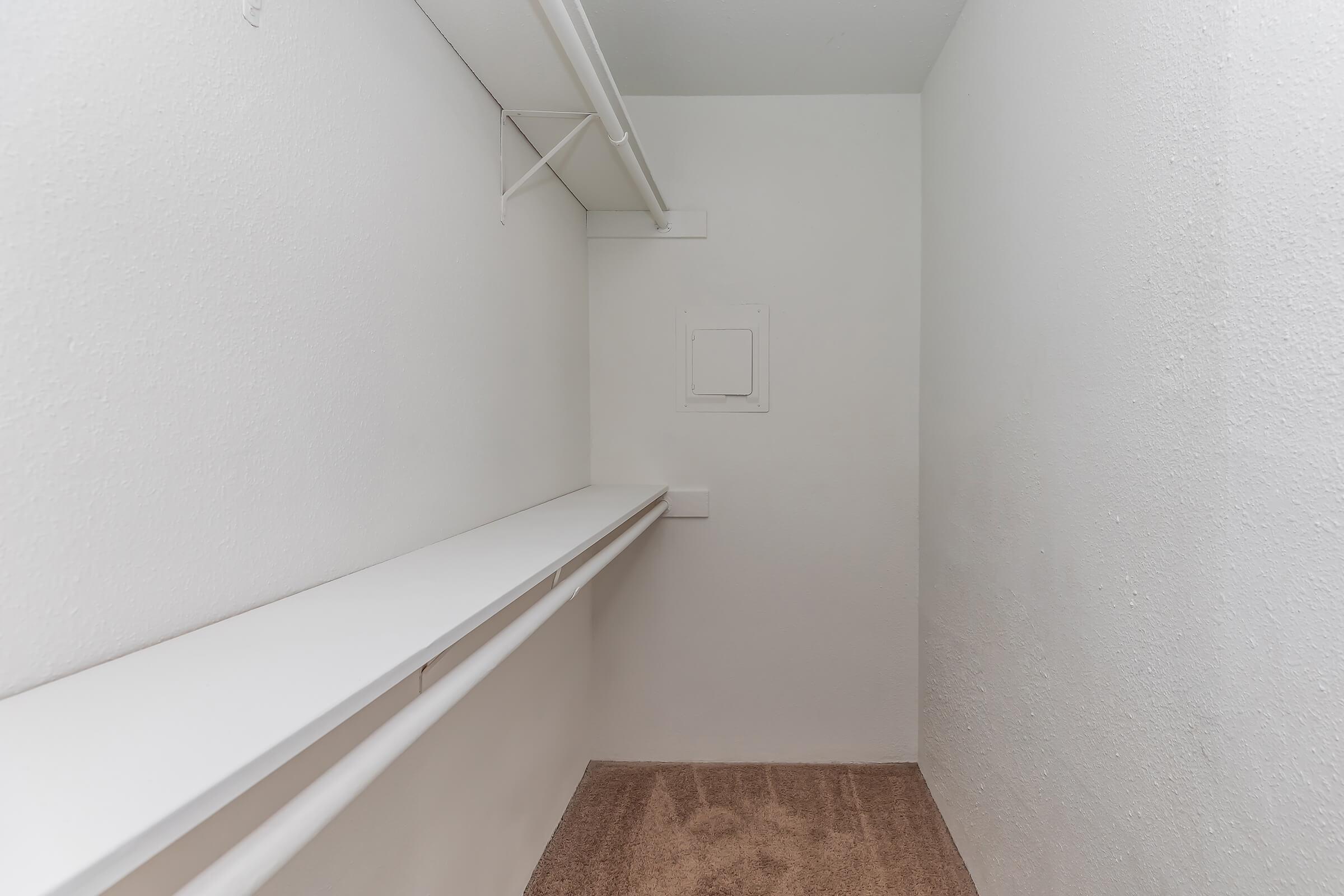 Empty closet with white walls and a bare wooden floor, featuring a long shelf on one side and a small panel on the back wall. The space is well-lit and has a minimalistic design.