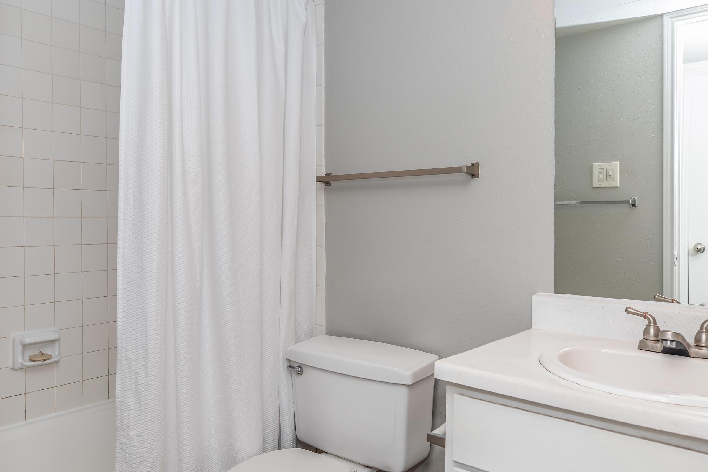 A clean bathroom featuring a white shower curtain, a bathtub with tiled walls, a white toilet, and a sink with a mirror above it. The walls are painted in a light gray color, and there is a towel rack mounted on the wall near the shower.