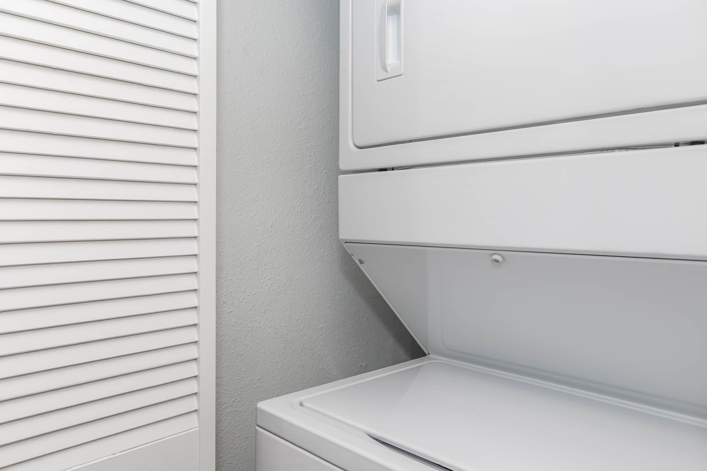 A corner of a laundry room featuring a stacked washer and dryer in white. A light gray wall is adjacent, and a closed white louvered door is visible, creating a clean and organized look.