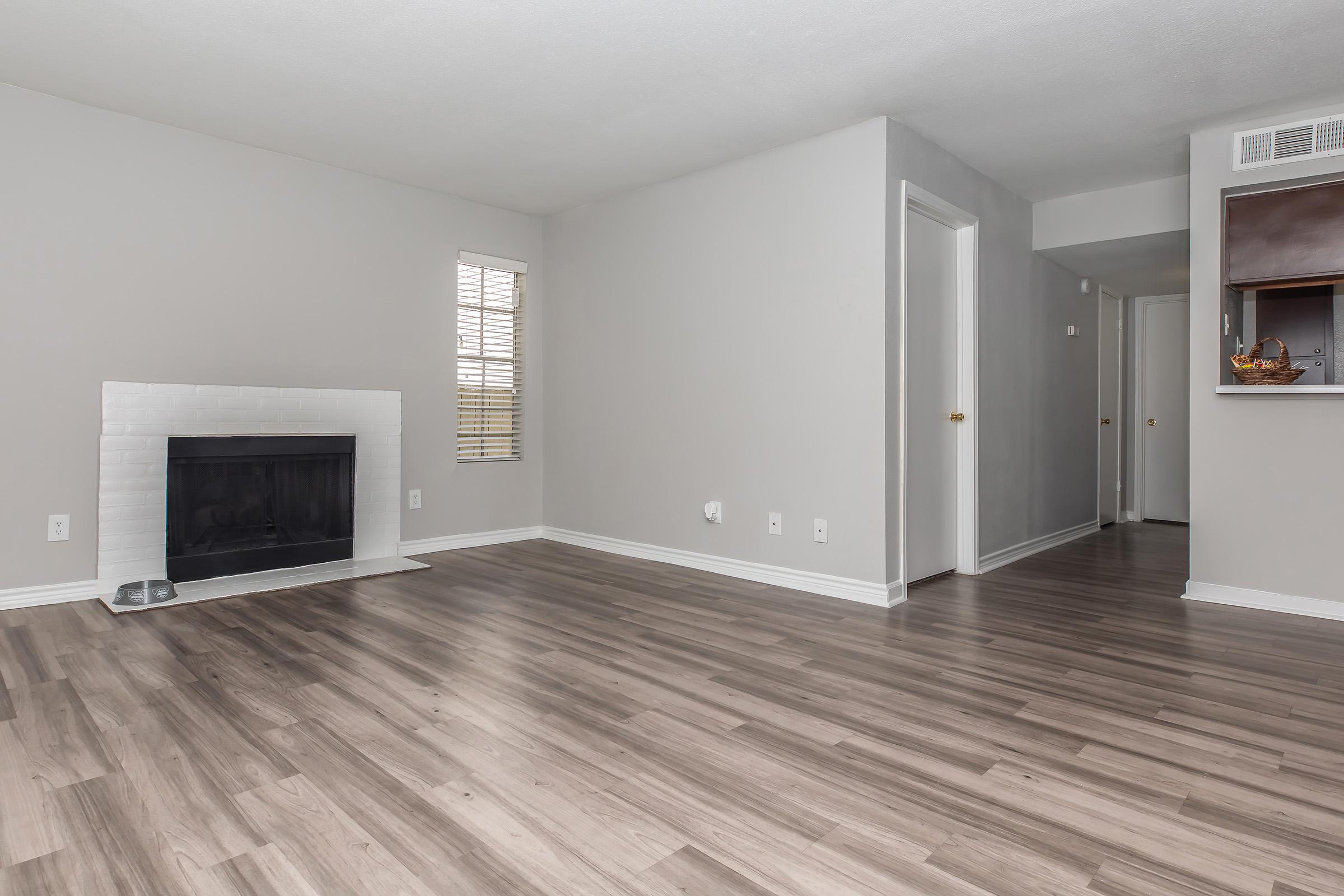 A spacious, empty living room featuring light-colored walls and modern wood-like flooring. There is a white brick fireplace on one side and a narrow window with horizontal blinds letting in natural light. A door leads to a hallway on the right side of the image.