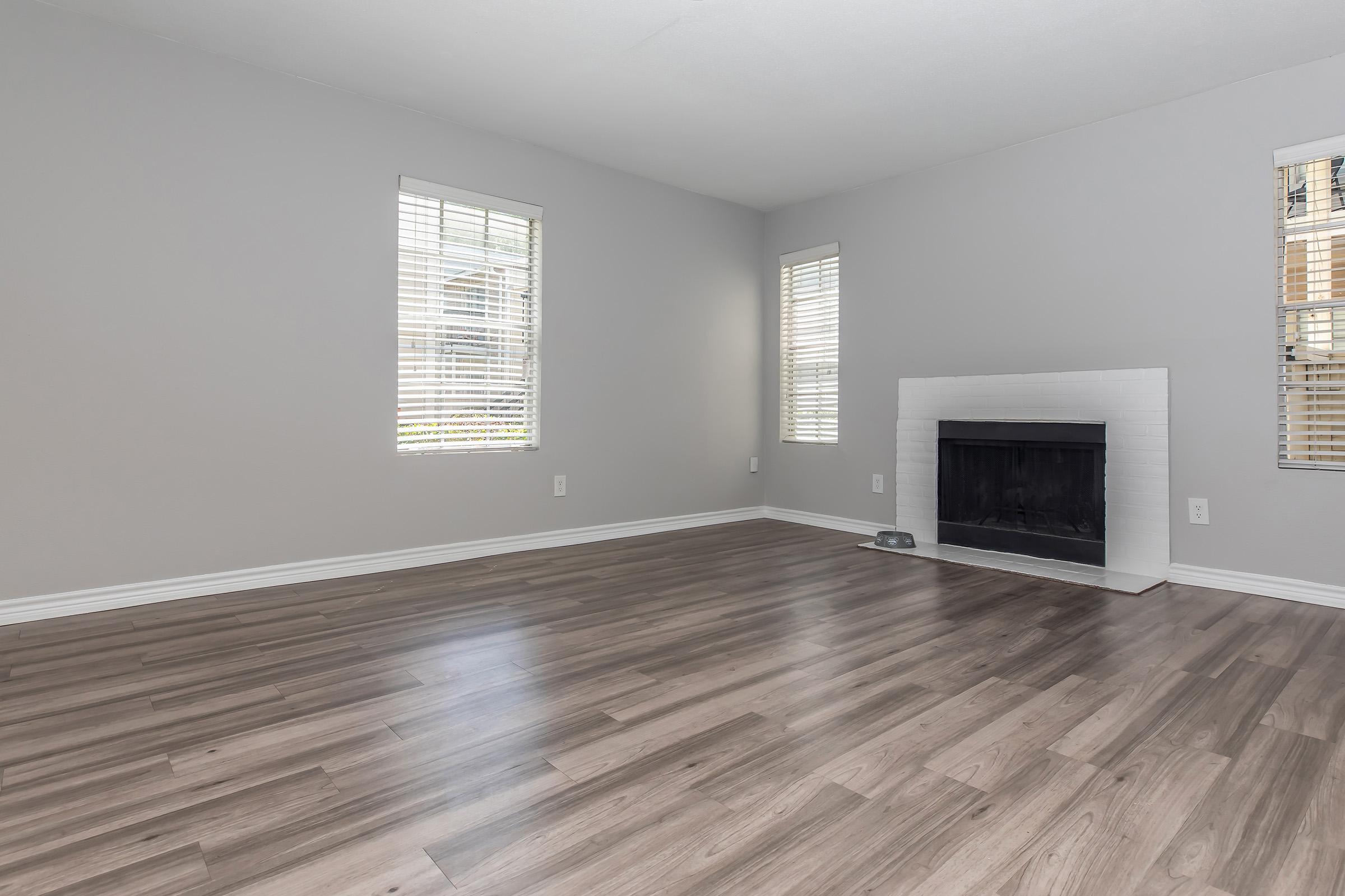 Empty living room with gray walls and light-colored wooden flooring. There are two windows with horizontal blinds allowing natural light to enter. A white tiled fireplace is situated against one wall, creating a focal point in the room. The overall ambiance is bright and spacious.