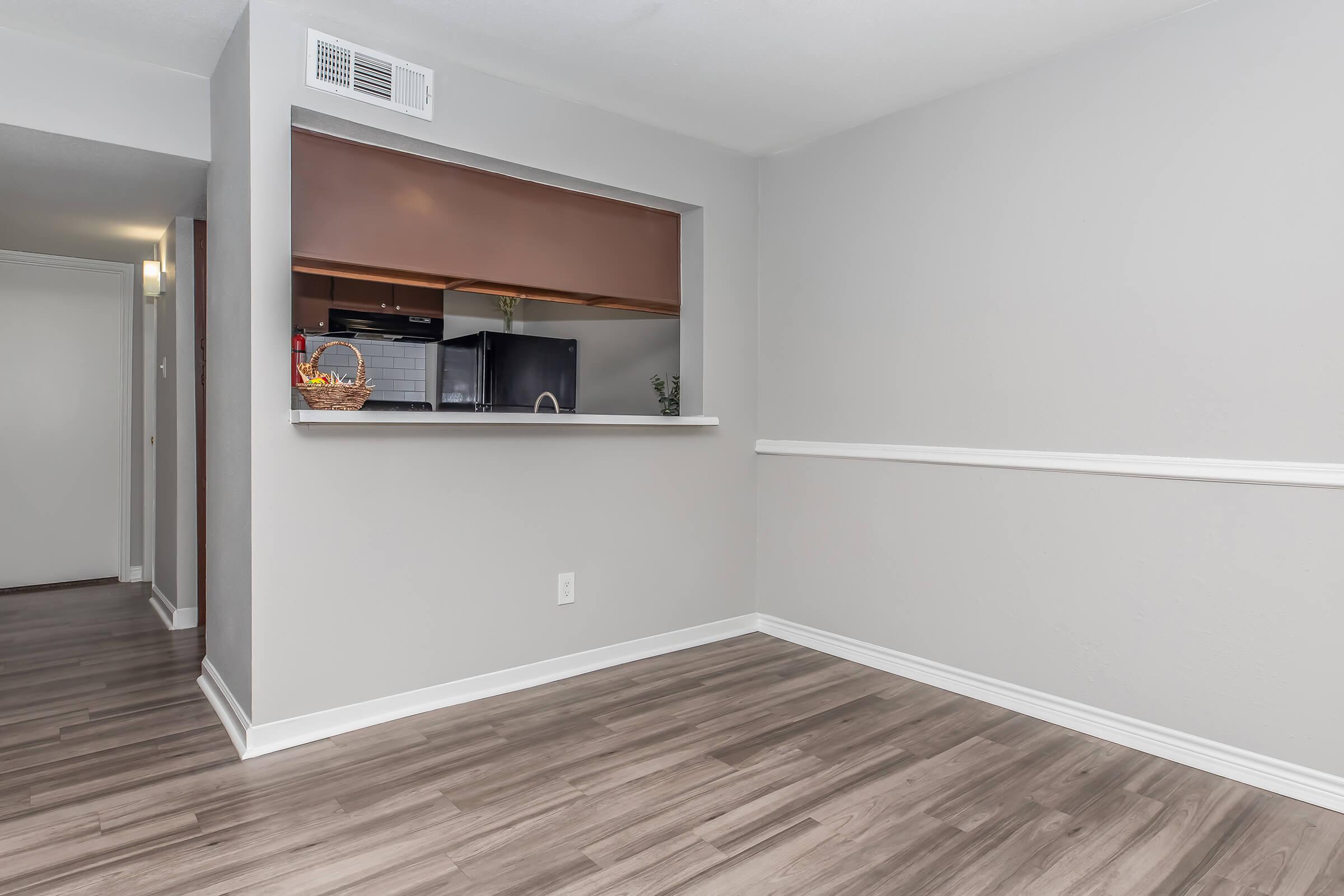 Interior view of a modern living space featuring light gray walls, a sectioned-off kitchen visible through an open window, and a wood-like floor. There is a small decorative basket on the countertop and a white chair rail along the wall, creating a clean and inviting atmosphere.