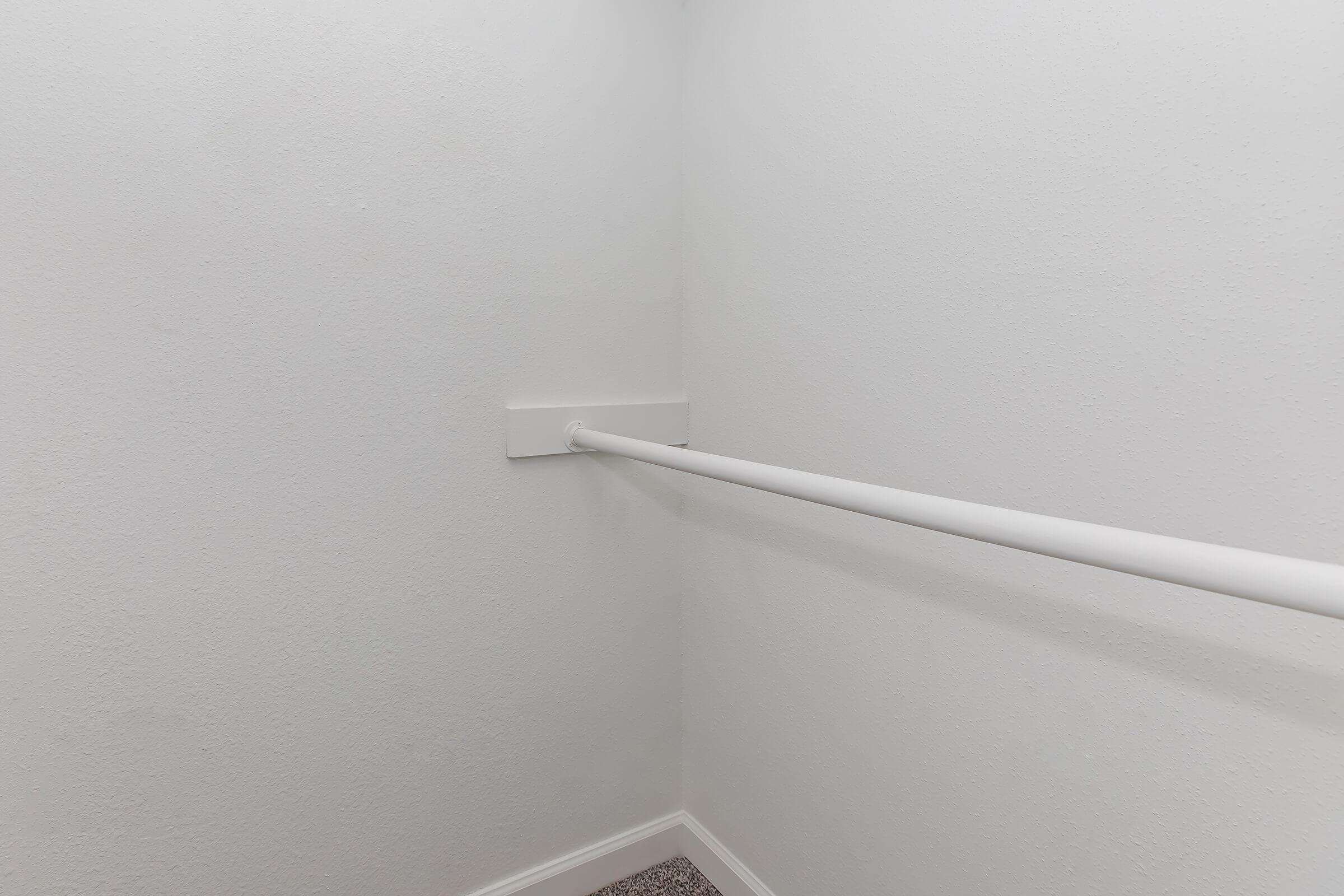 Empty closet corner with a white wall and a white clothing rod mounted along the wall. The floor has a light-colored carpet, and the overall space appears clean and unoccupied.