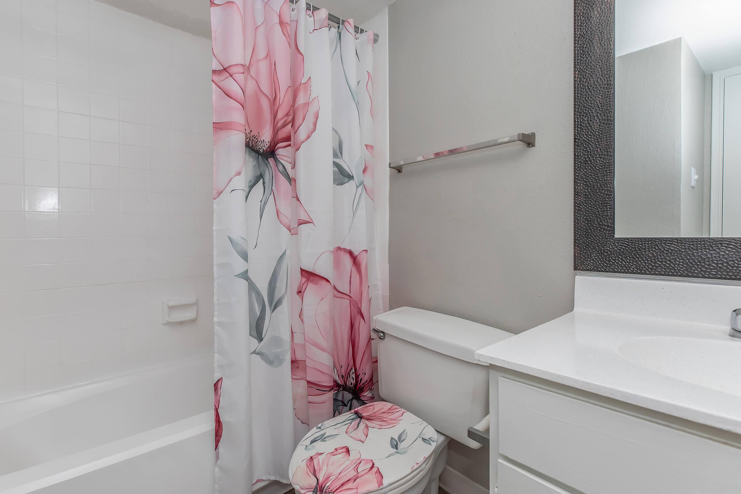 A bathroom featuring a bathtub with a white tile surround, a toilet, and a vanity. The shower curtain is adorned with large pink flowers against a white background, adding a vibrant touch to the space. A mirror above the vanity reflects the room's light, enhancing the brightness.