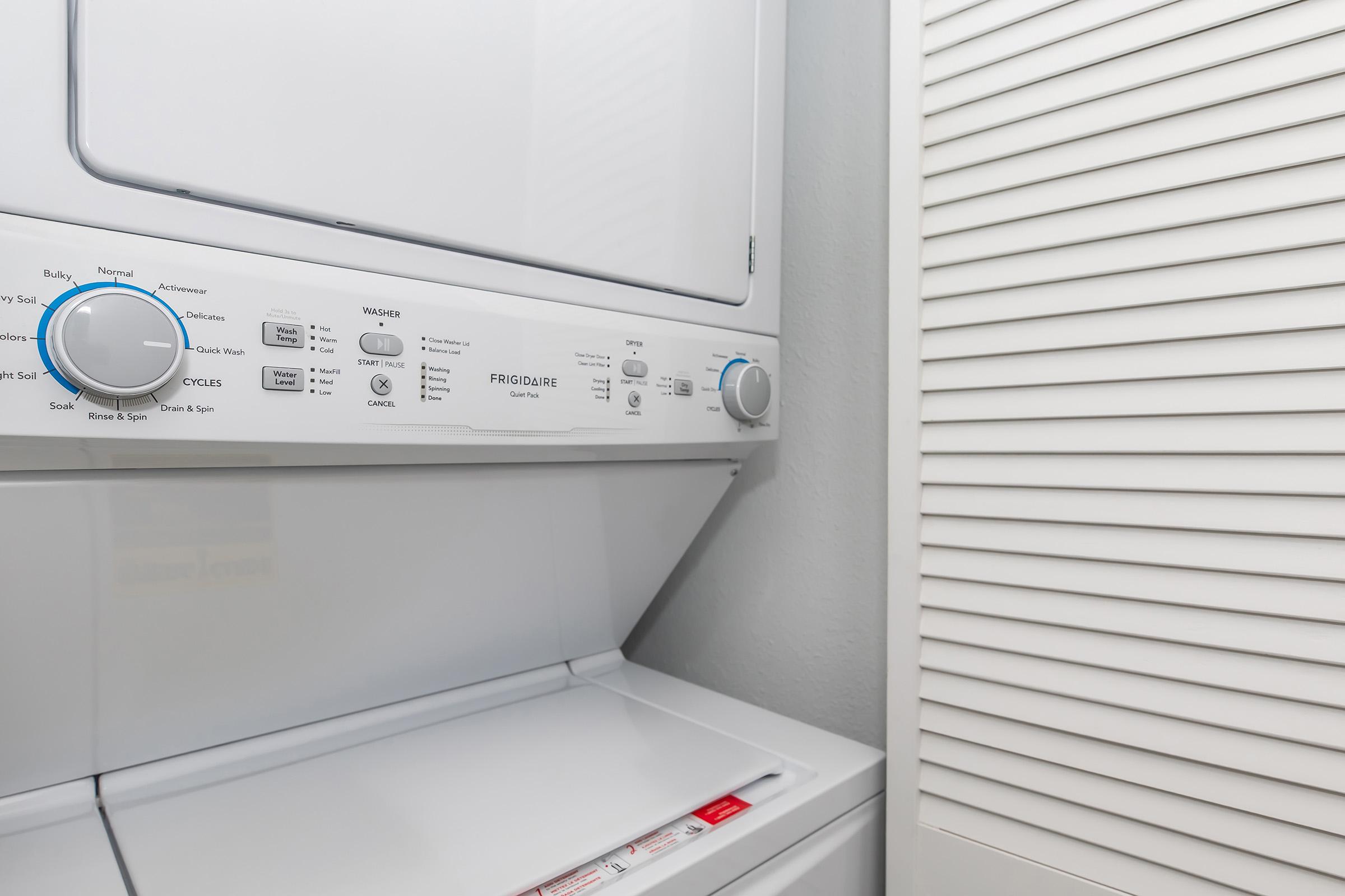 A stacked washer and dryer unit in a laundry area, with clear control panels featuring various settings and cycles. Nearby, a white louvered door suggests additional storage or space. The appliances are neatly arranged against a plain wall.