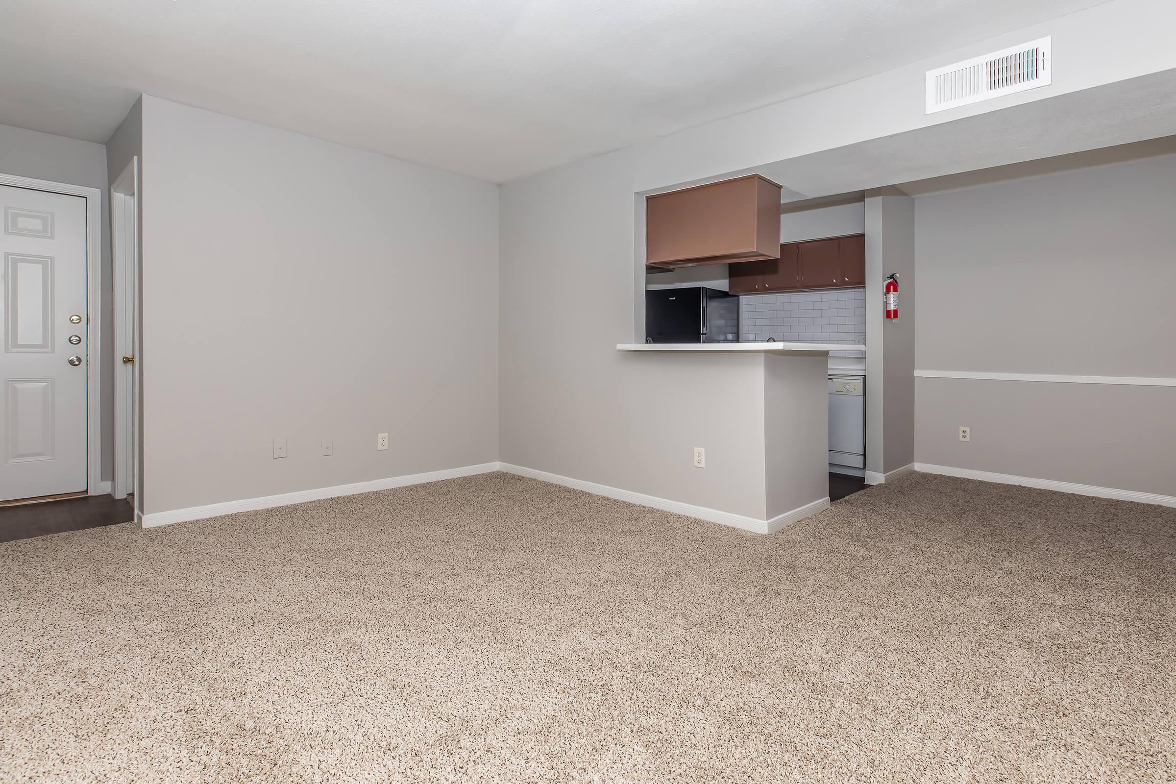 A spacious, neutral-colored living area with beige carpet and light gray walls. A doorway leads to another room, while an opening reveals a kitchen with brown cabinetry in the background. The space appears clean and ready for furnishing, creating a welcoming atmosphere.