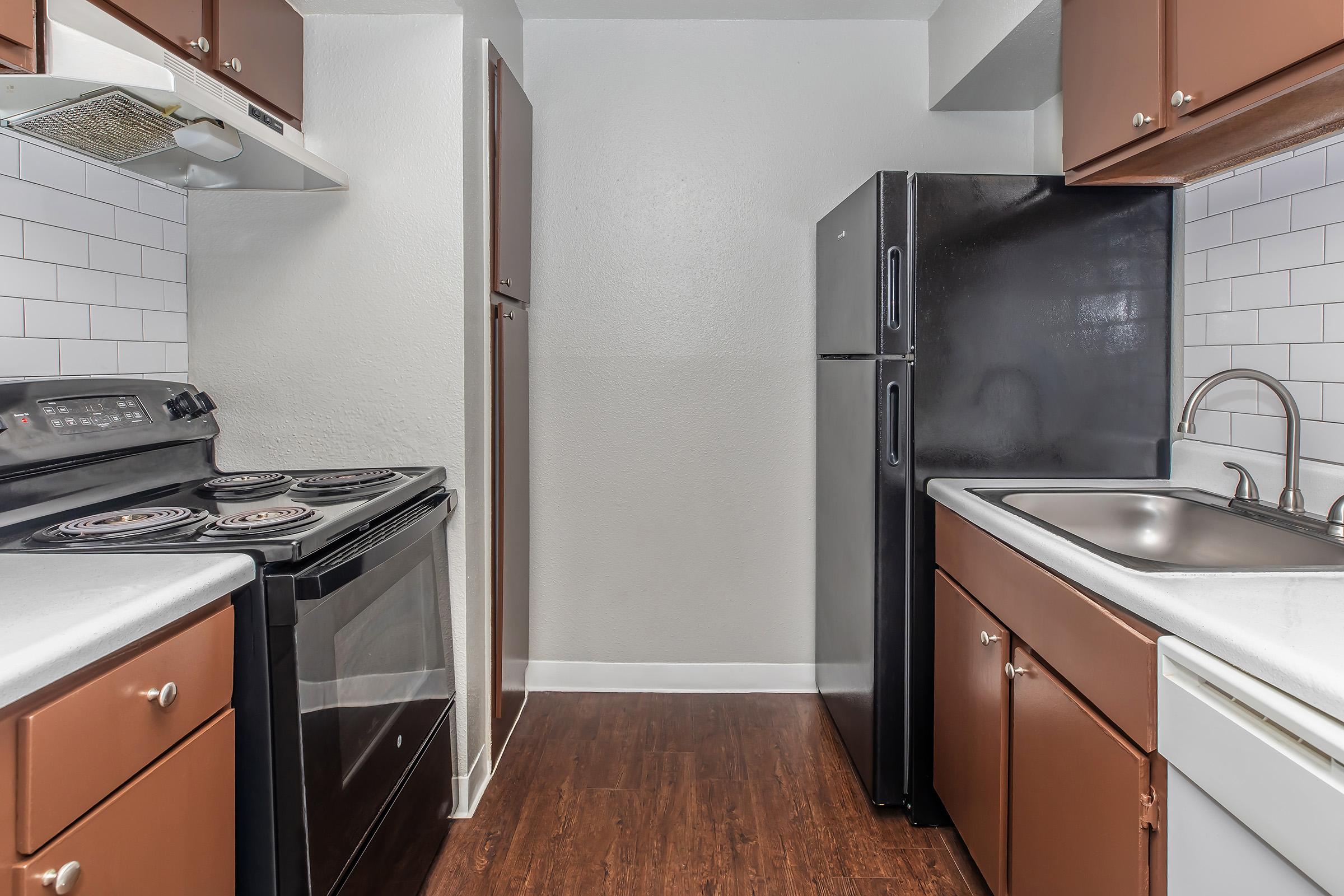 Modern kitchen featuring dark wood cabinets, a black refrigerator, and a black stove. The counters are light-colored, and there's a stainless steel sink. White subway tile backsplash adds a contemporary touch. The floor is dark wood, contributing to the overall warm aesthetic of the space.