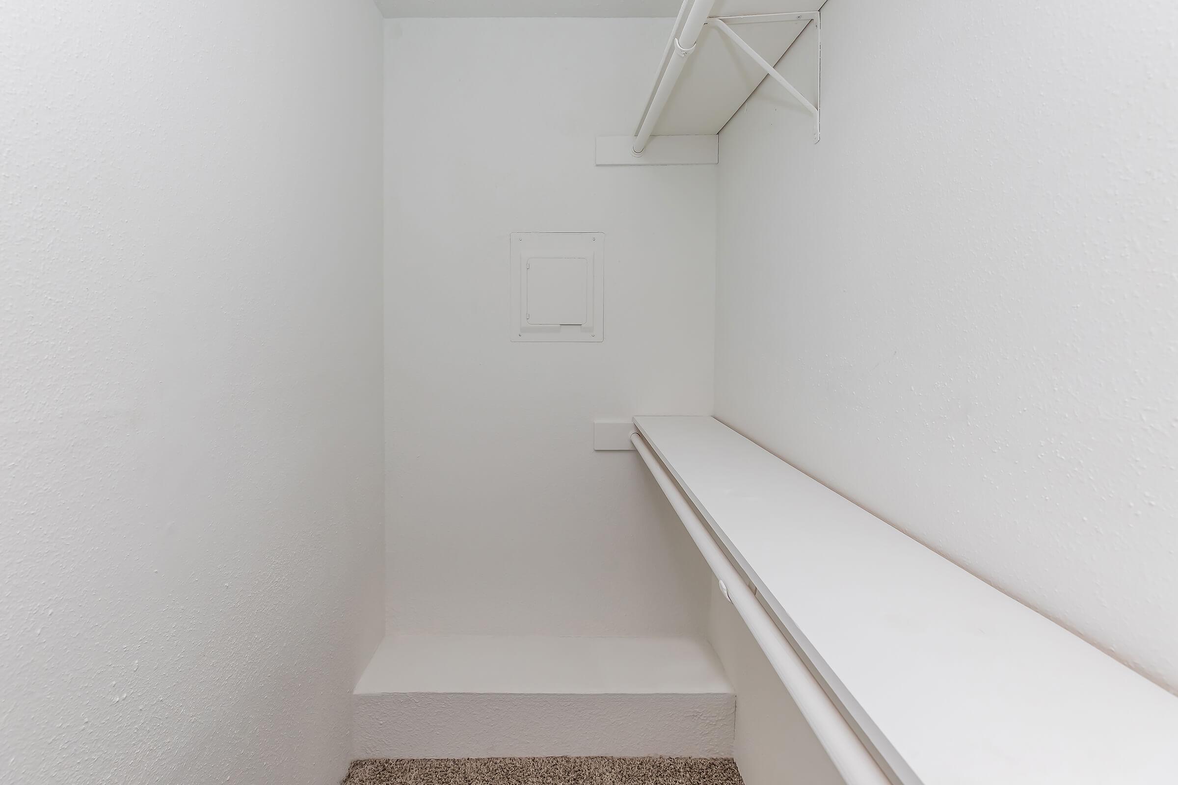 A clean, empty closet with white walls and a beige carpet. There is a long shelf along one side, and a small access panel is visible on the wall. The space is well-lit and shows no items or decorations, giving it a simple and minimalistic appearance.