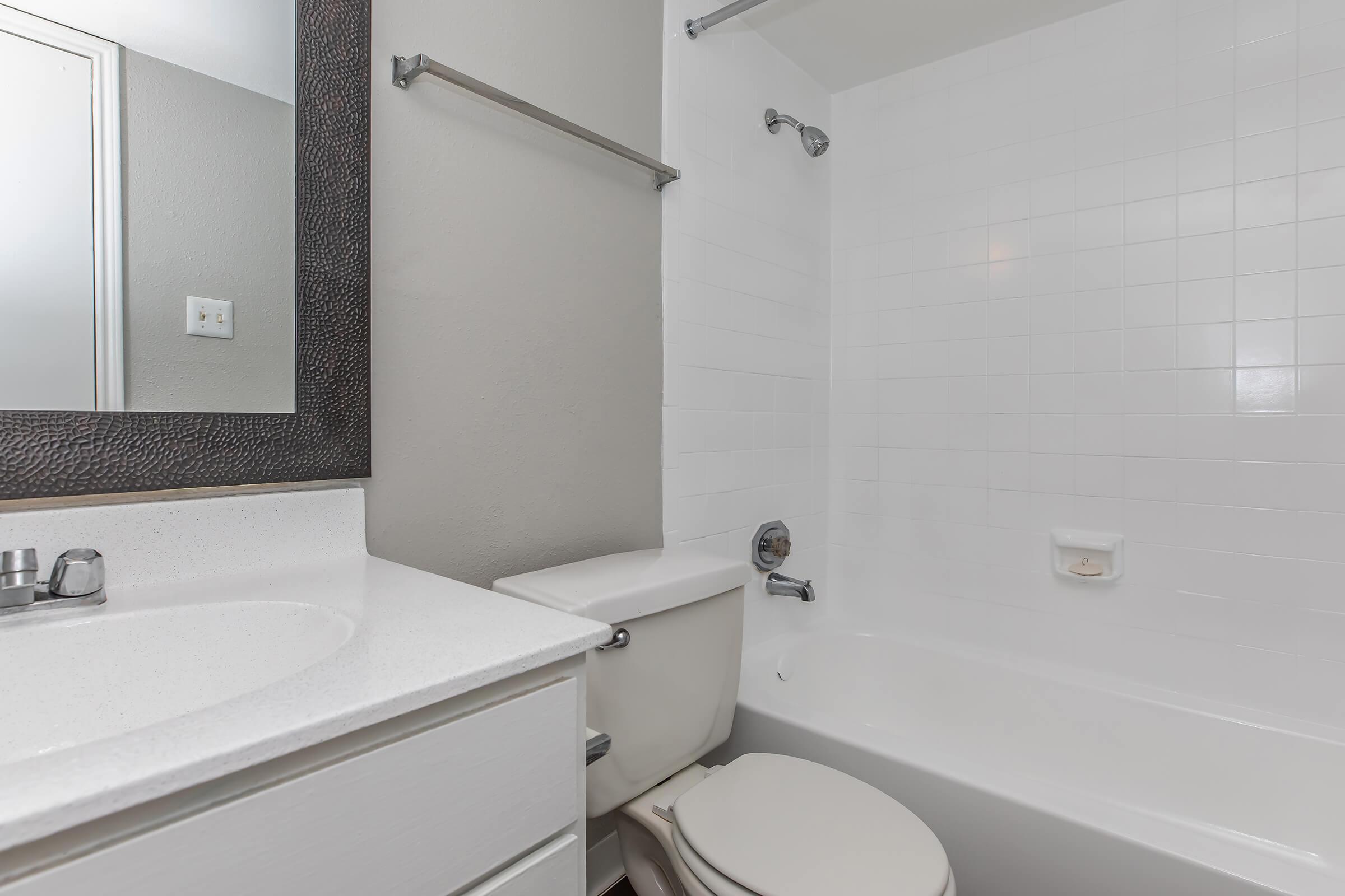 A clean and modern bathroom featuring a white tiled bathtub, a toilet, and a large mirror above a white sink. The walls are painted in a light color, and there is a towel bar mounted on the wall. The overall design is simple and contemporary with minimal decor.