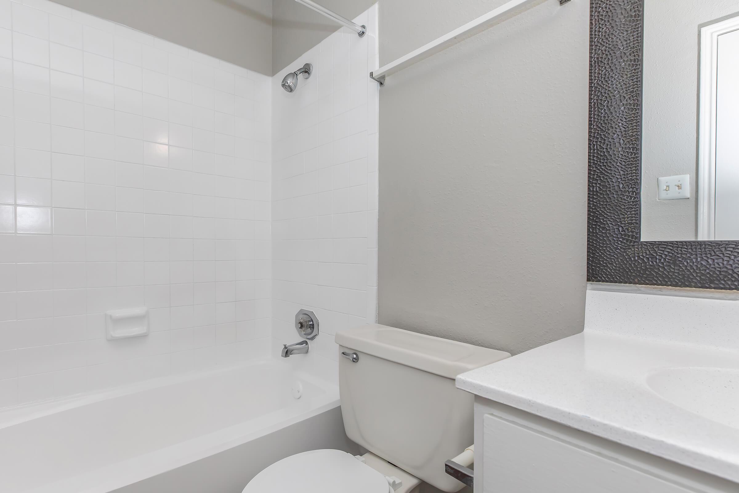 A clean, modern bathroom featuring a white tiled bathtub and shower, a white toilet, and a simple countertop with a mirror above it. The walls are painted in a neutral color, providing a bright and airy atmosphere.