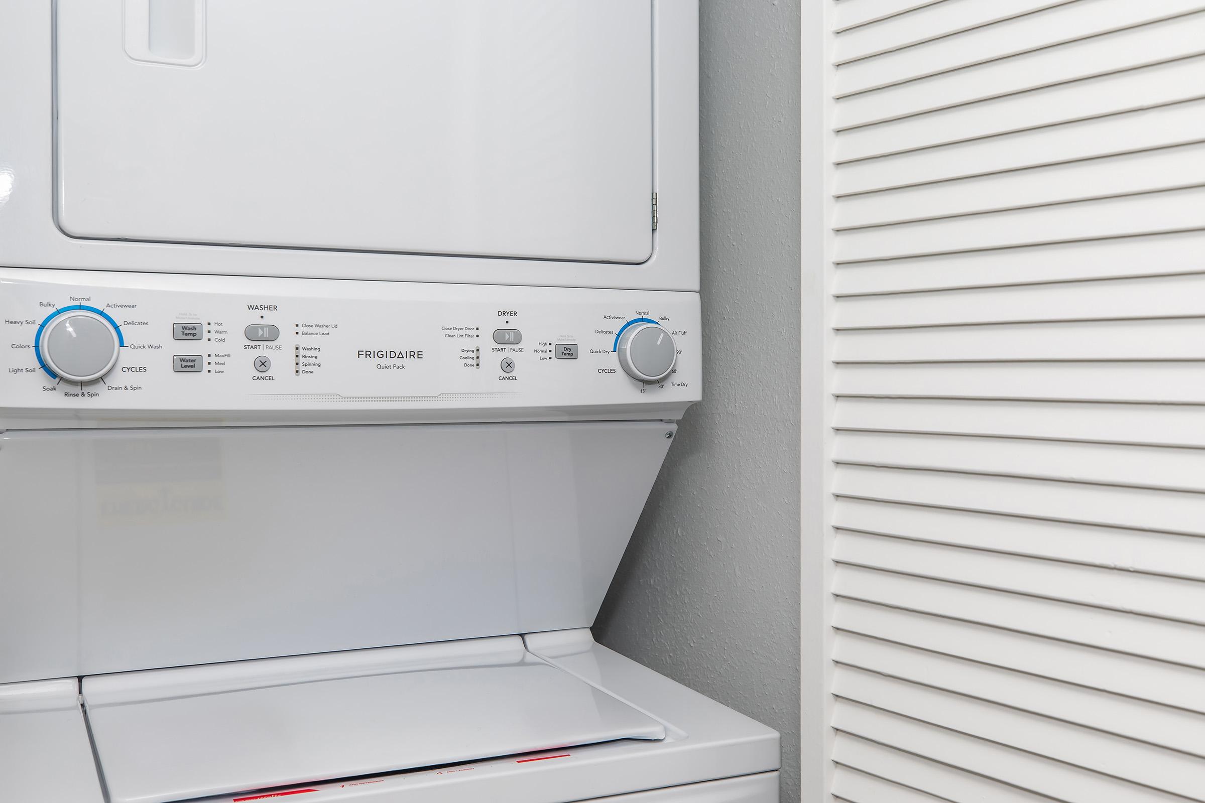 A stacked washer and dryer unit by Frigidaire, featuring control knobs and buttons for various settings. The appliance is set against a gray wall with a closed louvered door on the right.
