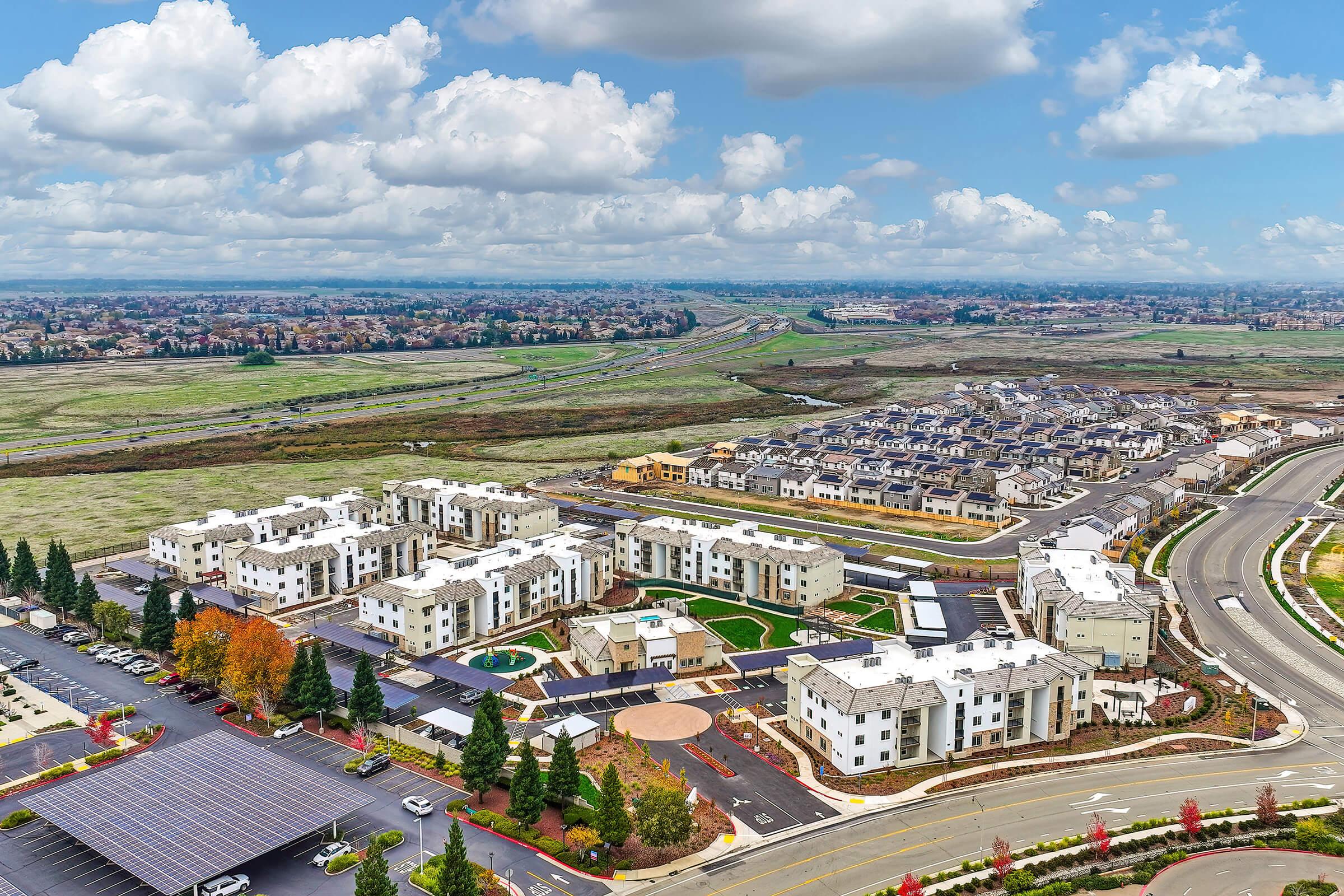 Aerial view of a residential area featuring modern multi-family buildings, landscaped gardens, and parking spaces. In the background, there are open fields and distant suburban developments under a partly cloudy sky. Roads and pathways connect the buildings, creating a well-planned community layout.