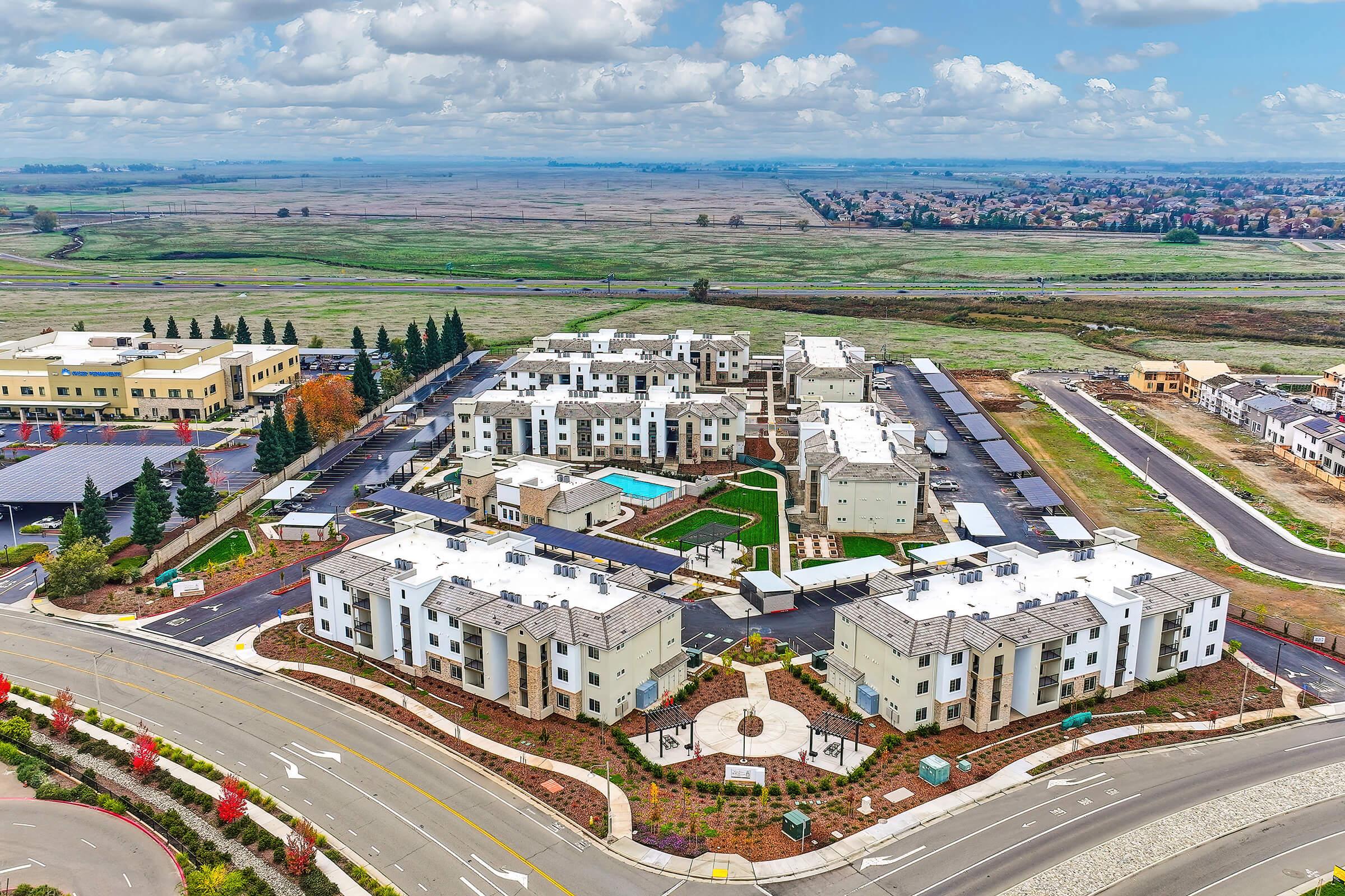 Aerial view of a modern residential complex with multiple buildings featuring green landscaping, a swimming pool, and parking areas. Surrounding fields and scattered homes are visible in the background under a partly cloudy sky.