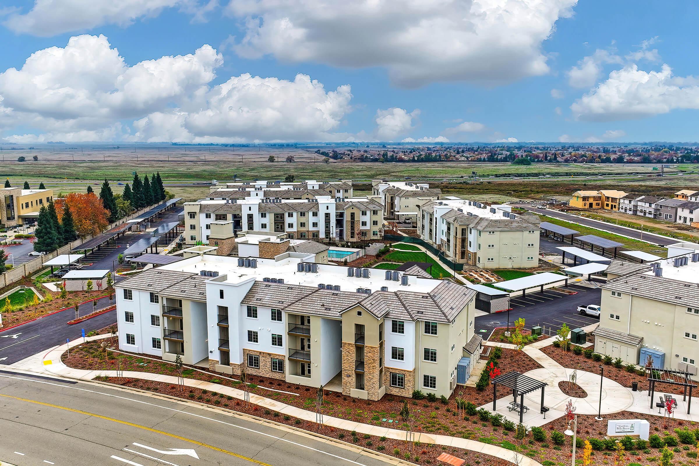Aerial view of a modern apartment complex featuring multiple buildings with white and beige exteriors, landscaped areas, and a parking lot. In the background, open fields and a cloudy sky can be seen, creating a serene suburban atmosphere.