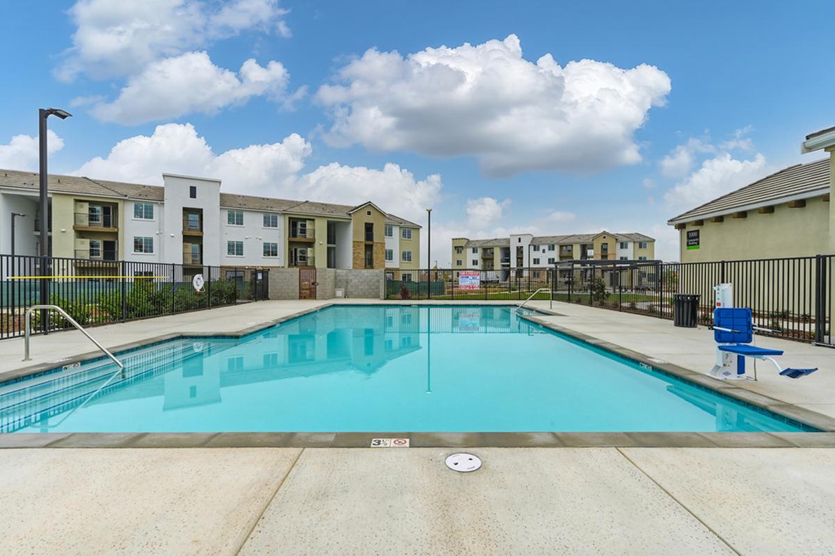 A clear swimming pool surrounded by a protective fence, with lounge chairs nearby. In the background, there are modern residential buildings with various colors under a bright blue sky with scattered clouds.