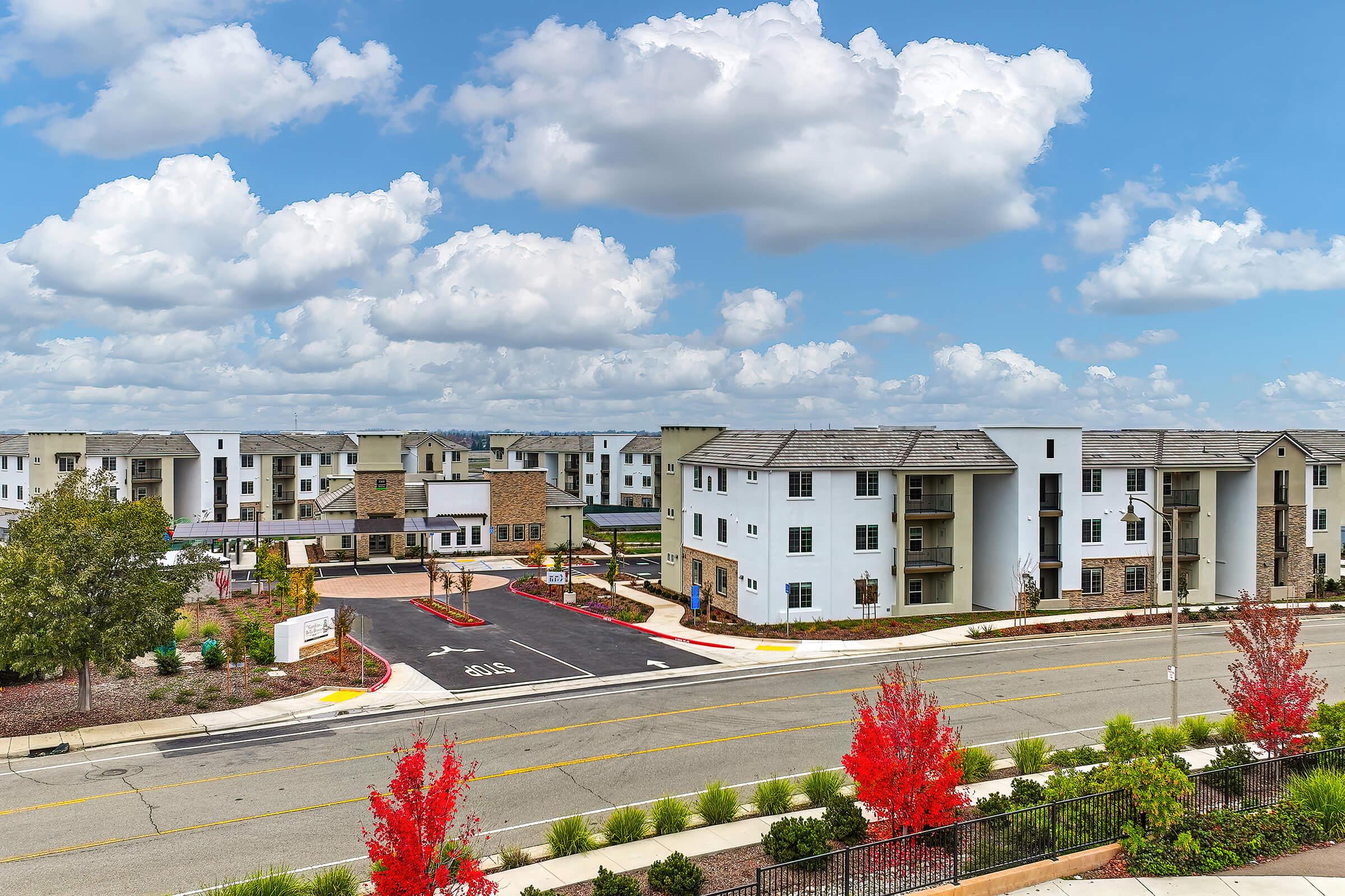 A modern residential complex featuring multiple buildings with a mix of white and stone walls. The area includes landscaped pathways, blooming red foliage, and a clear blue sky with fluffy clouds. The image captures a welcoming community atmosphere with ample parking and greenery.