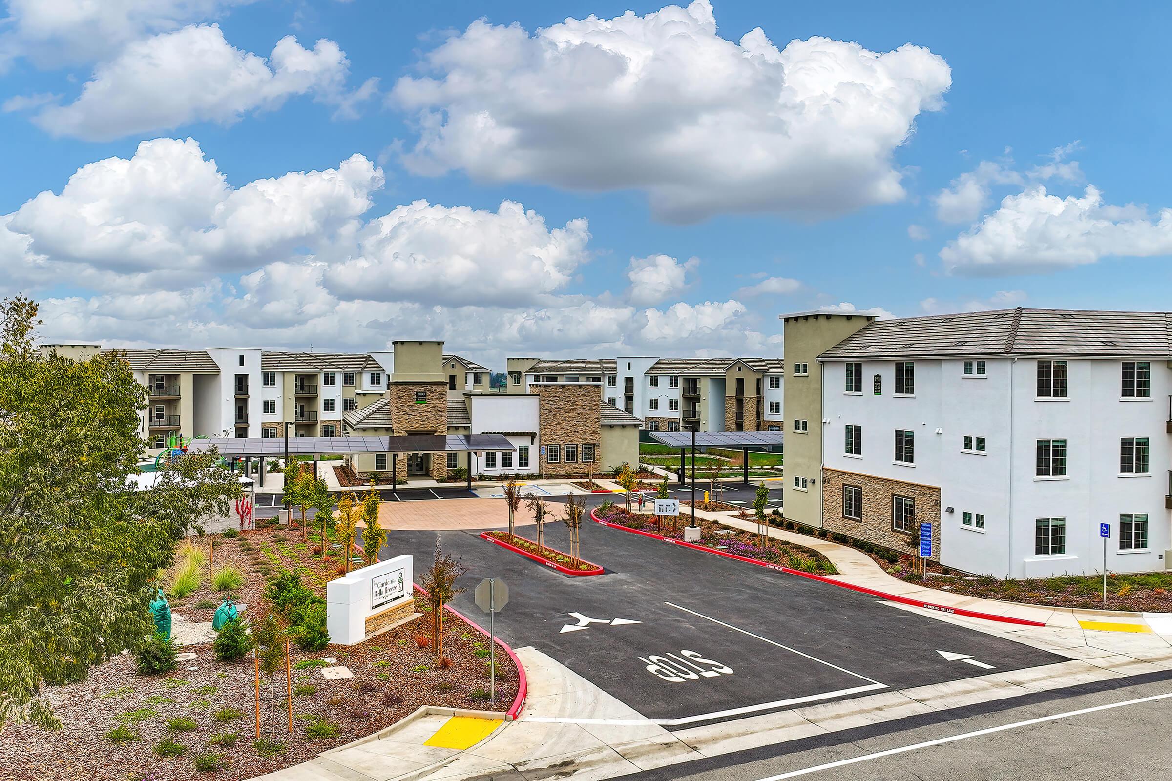 A modern residential complex featuring several multi-story buildings with a mix of white and stone facades. The entrance includes a driveway and landscaped areas with trees and plants. The sky is bright with fluffy clouds, creating a welcoming atmosphere.