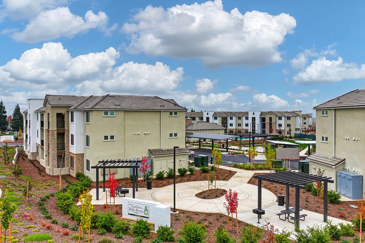 A well-maintained residential complex featuring multiple modern buildings surrounded by landscaped green areas, seating structures, and pathways. The sky is partly cloudy, creating a bright atmosphere. Facilities and amenities are evident, indicative of a community living environment.