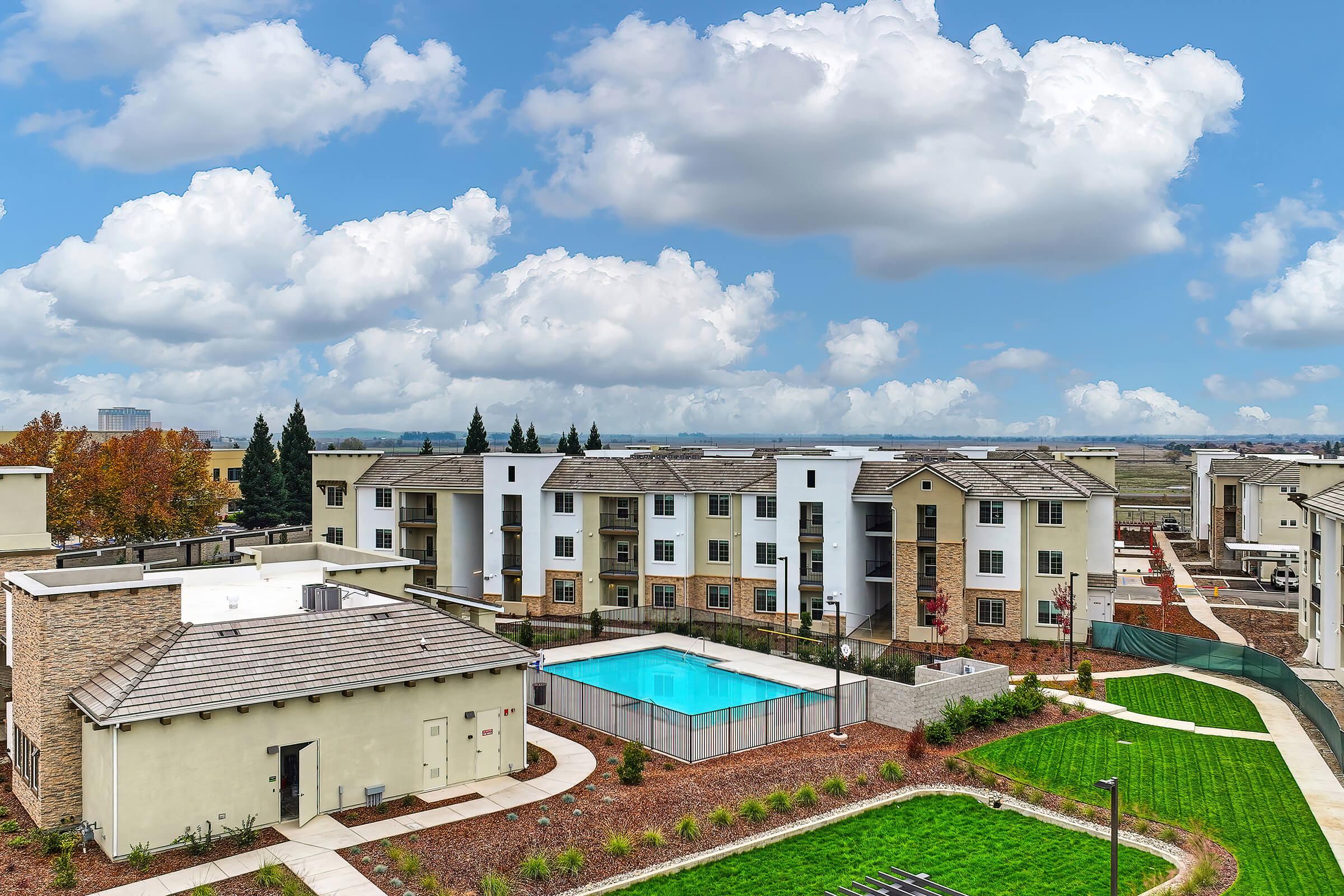 Aerial view of a residential complex featuring modern apartment buildings arranged around a central pool area. Lush green landscaping surrounds the buildings, with walkways and seating areas. Fluffy clouds fill the blue sky in the background, enhancing the inviting atmosphere of the community.