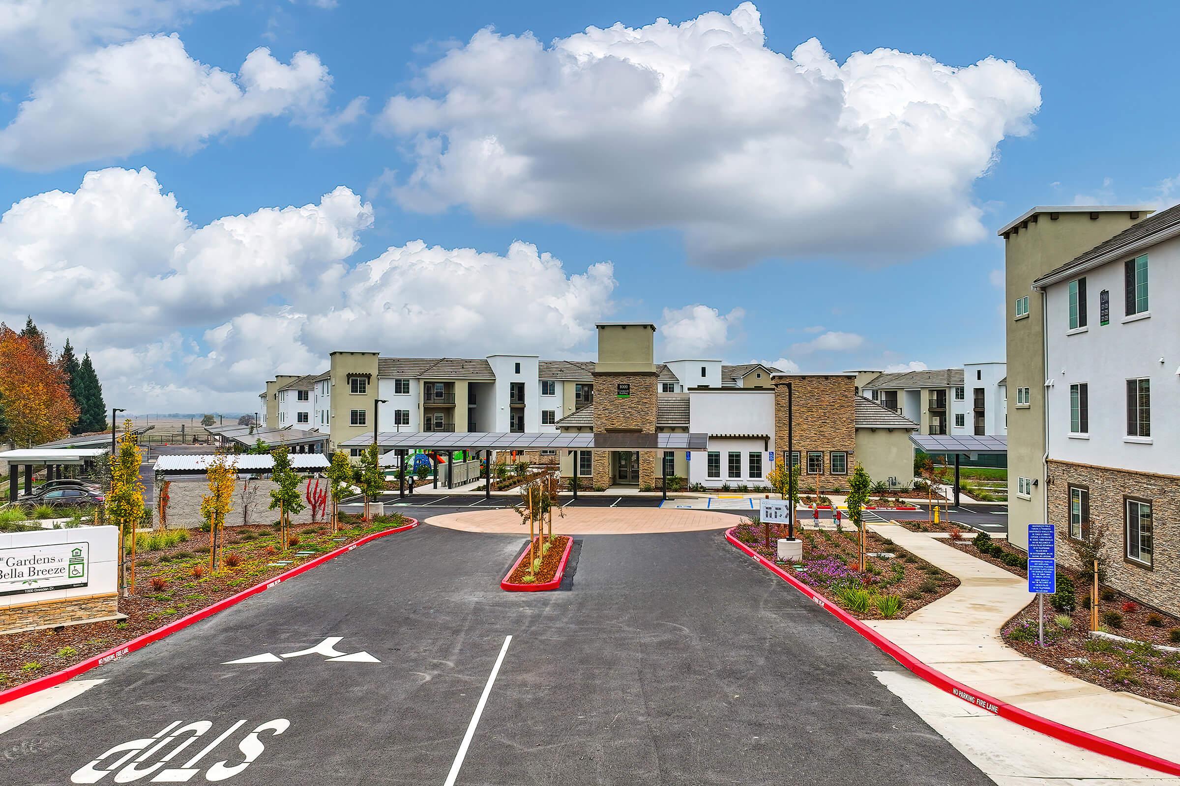A modern multi-story residential building with a stone façade and landscaped grounds. The entrance features a wide driveway, signage indicating a stop, and decorative plants. It is a sunny day with fluffy clouds in the blue sky, creating a welcoming atmosphere for residents and visitors.
