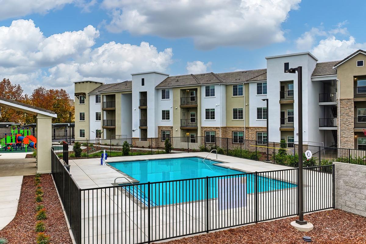 A view of an apartment complex featuring a swimming pool in the foreground. The pool is enclosed by a black fence, and there are landscaped areas with shrubs around it. In the background, several multi-story residential buildings are visible under a partially cloudy sky.