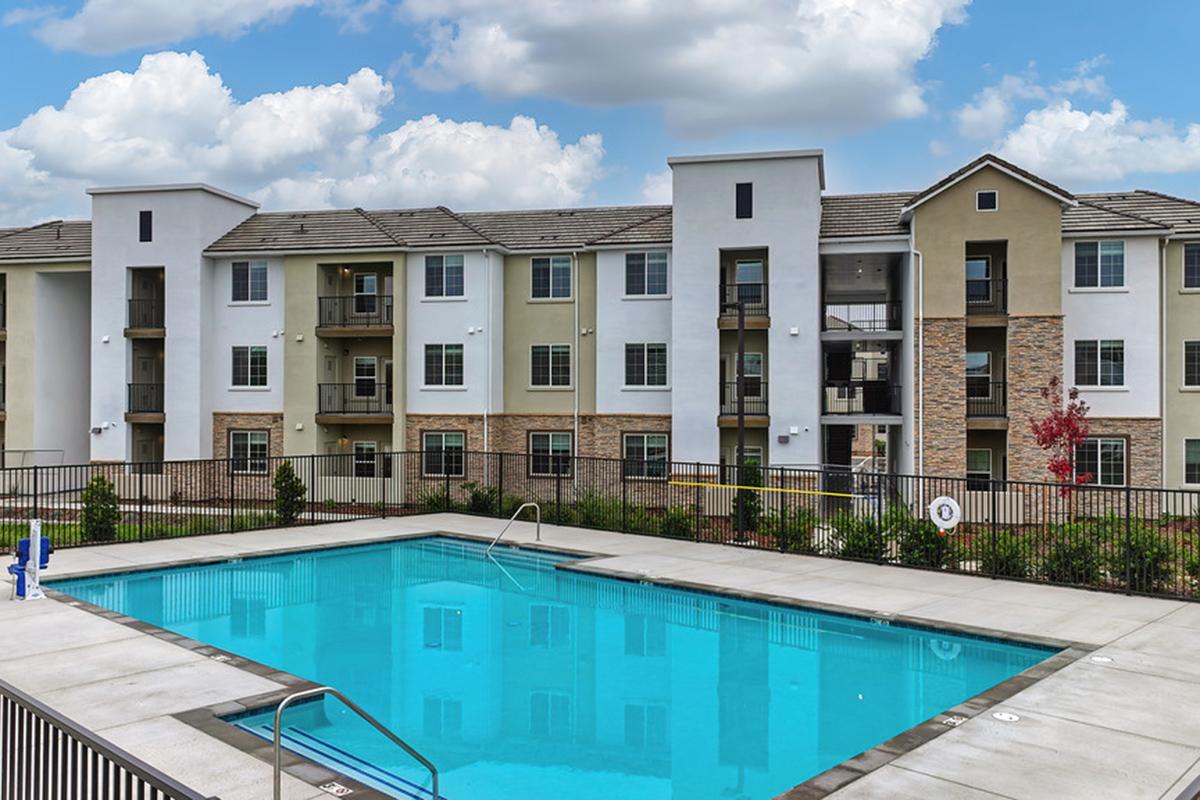 A modern apartment building with a swimming pool in the foreground. The pool area features lounge chairs and a surrounding fence. The building has a mix of light-colored stucco and brick, with several balconies visible. The sky is partly cloudy, creating a bright atmosphere.