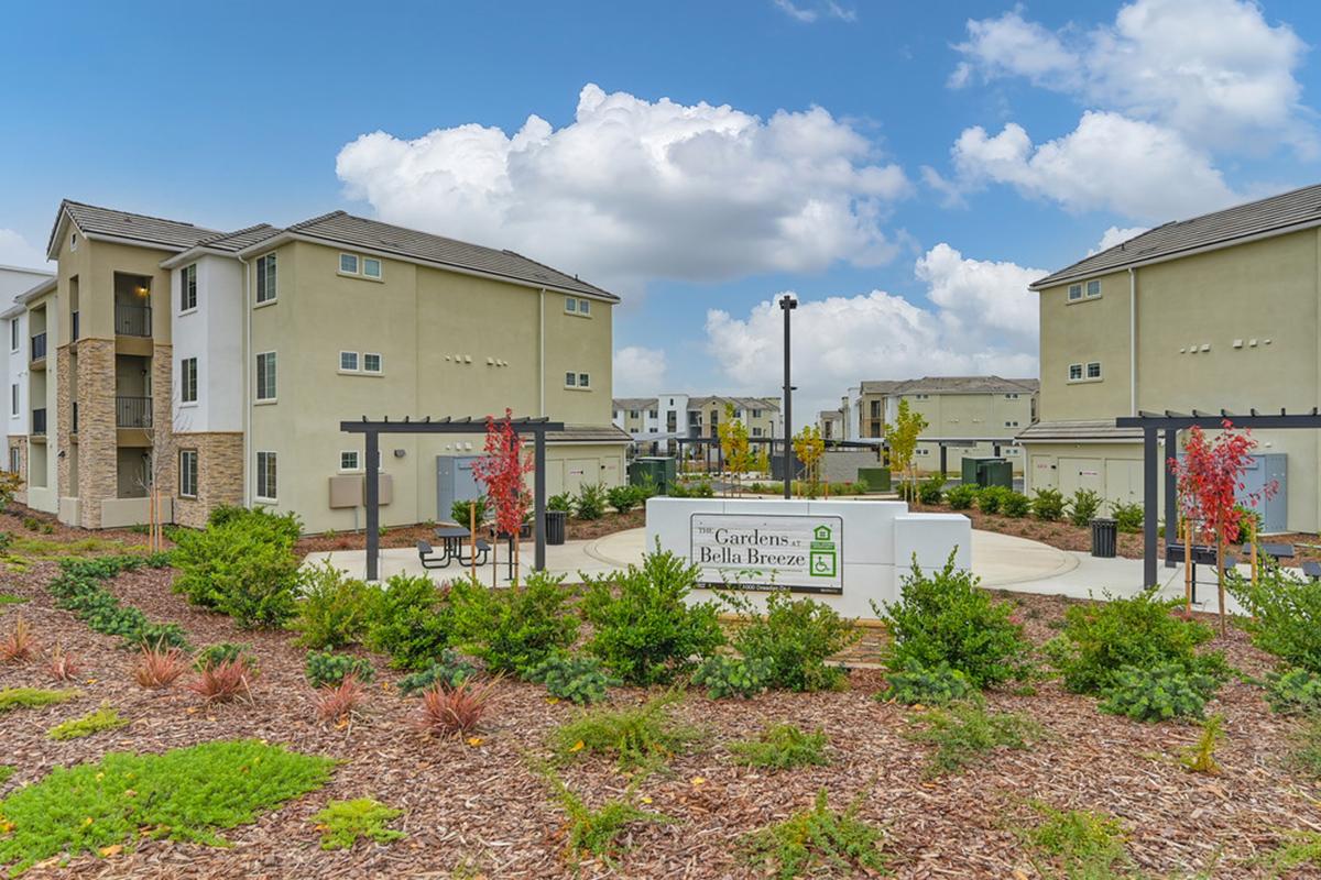 Modern apartment complex featuring multiple three-story buildings surrounded by landscaped grounds with shrubs and flowers. The entrance sign reads "Gardens at Bella Breeze." A central courtyard area with seating is visible under a partly cloudy sky.