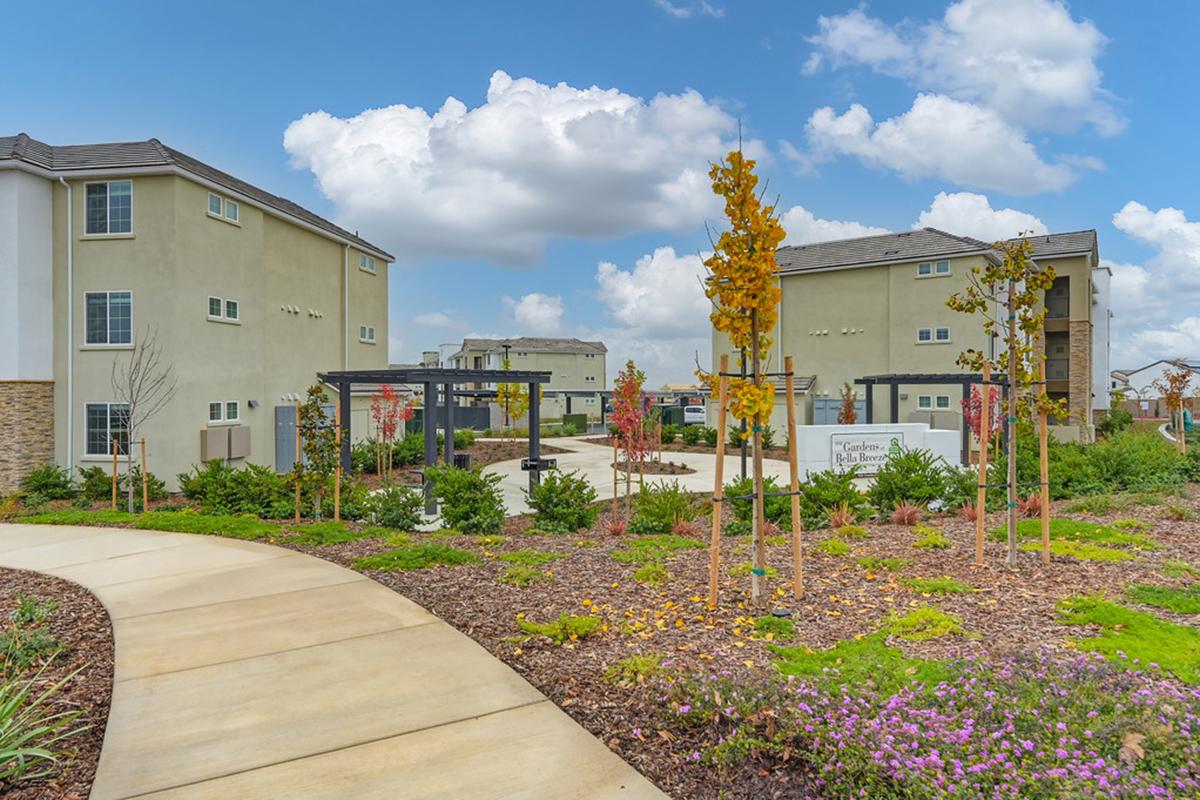 A landscaped courtyard featuring a pathway, modern multi-story residential buildings, and decorative plants. The sky is partly cloudy, creating a bright atmosphere. The area includes seating and communal spaces, inviting relaxation and outdoor activities.