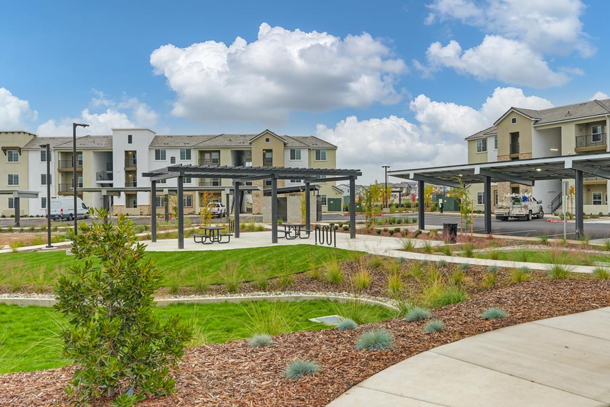 A modern residential complex featuring landscaped green spaces, picnic areas with shaded structures, and nearby parked vehicles. Light-colored buildings with multiple stories are visible in the background, under a partly cloudy blue sky.
