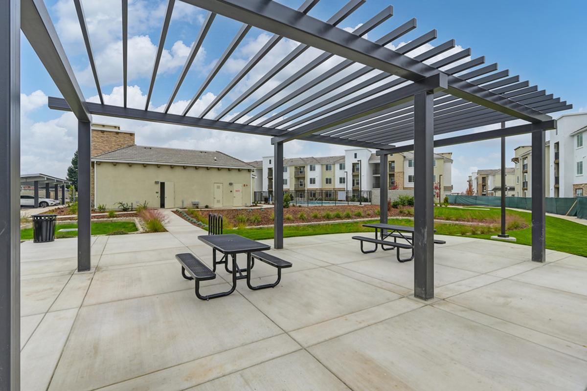 A modern outdoor seating area featuring a shaded pergola with picnic tables, surrounded by landscaped grounds and multi-story residential buildings under a partly cloudy sky.