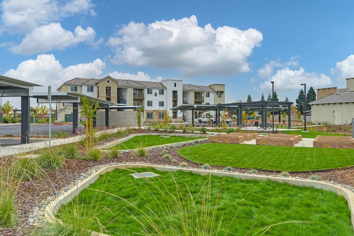 A landscaped outdoor area featuring green lawns, decorative stone pathways, and modern residential buildings in the background. The space includes shaded seating areas under metal canopies, surrounded by trees and shrubs, under a blue sky with fluffy clouds.
