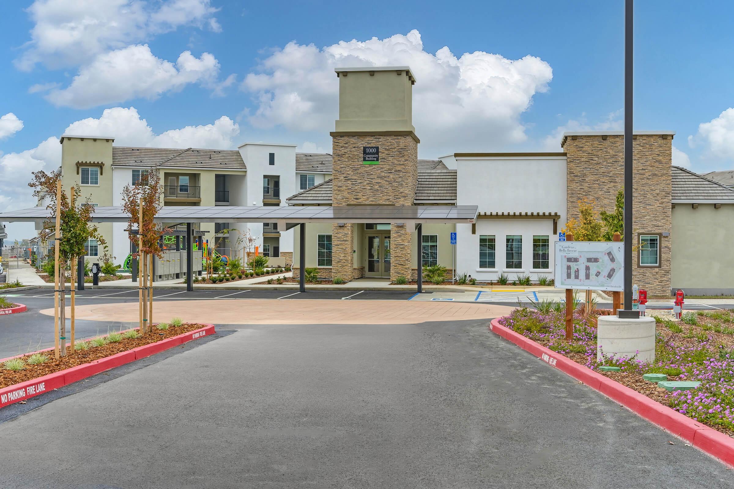 A modern building with a stone façade and tall entrance is seen, featuring a paved driveway surrounded by landscaped gardens. There are parking spaces in front, and the sky is bright with fluffy clouds. The structure includes various windows and architectural details, showcasing a welcoming atmosphere.
