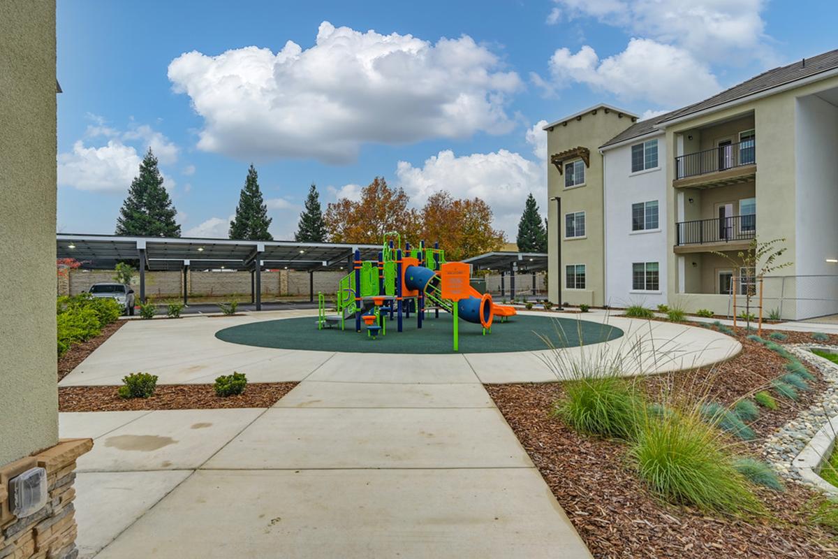 Playground area with colorful play structures surrounded by a green rubber surface and landscaped gardens. In the background, there are modern apartment buildings and shaded carports. Trees with autumn foliage provide a natural backdrop under a cloudy sky.