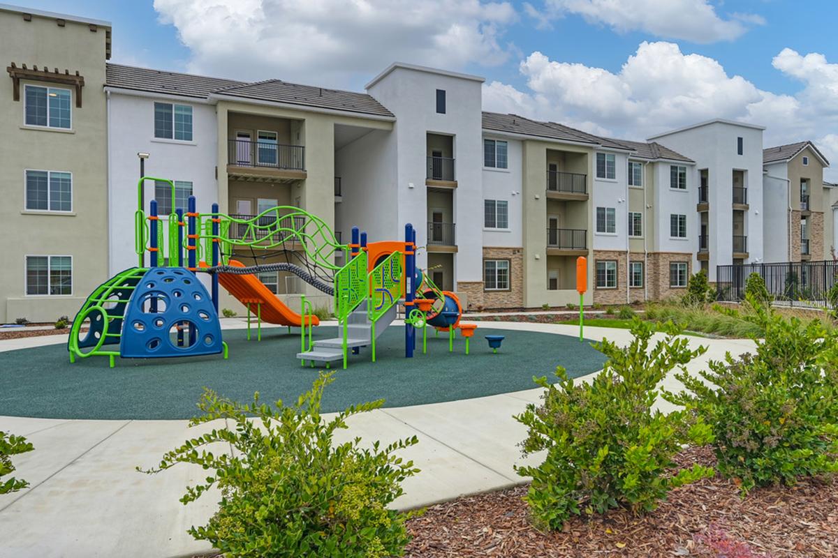 A vibrant children's playground featuring colorful equipment, including slides and climbing structures, surrounded by green landscaping and modern apartment buildings under a partly cloudy sky.