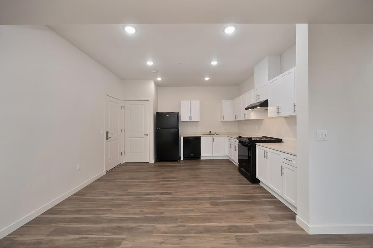 Modern kitchen in a newly designed apartment featuring white cabinets, black appliances, and laminate flooring. The open layout includes a stove, refrigerator, and ample counter space, with bright lighting illuminating the area.