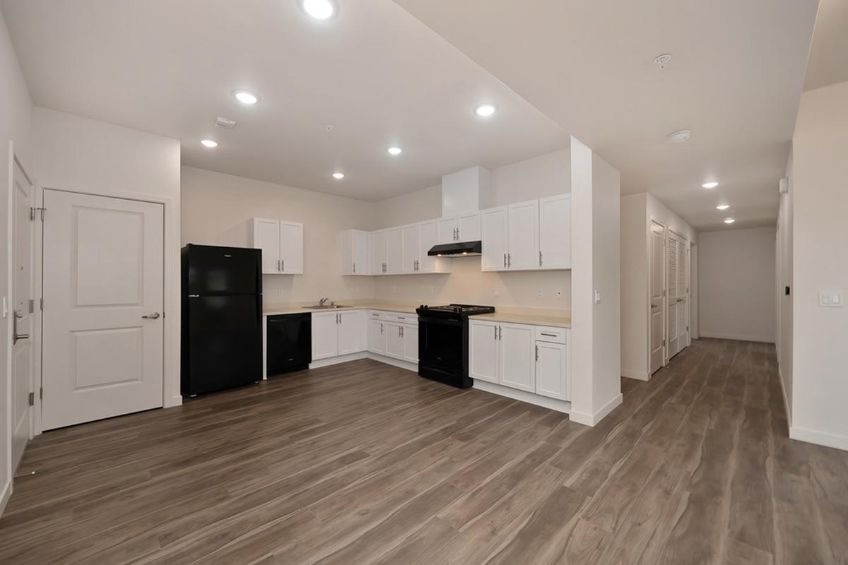 Modern kitchen featuring white cabinets, black appliances including a refrigerator and stove, and a spacious layout with wood-like flooring. Ample lighting from recessed fixtures enhances the bright and open atmosphere. A hallway to the right leads to additional rooms.