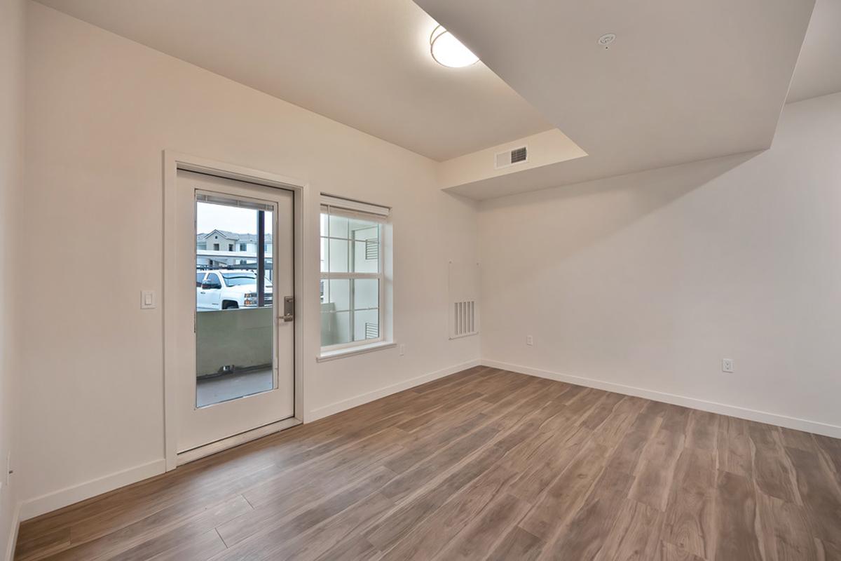 Empty room with wooden flooring, featuring a glass door leading to a balcony. A window is present on the side, allowing natural light to enter. The walls are painted white, and the ceiling is plain with a central light fixture. The overall appearance is modern and minimalistic.