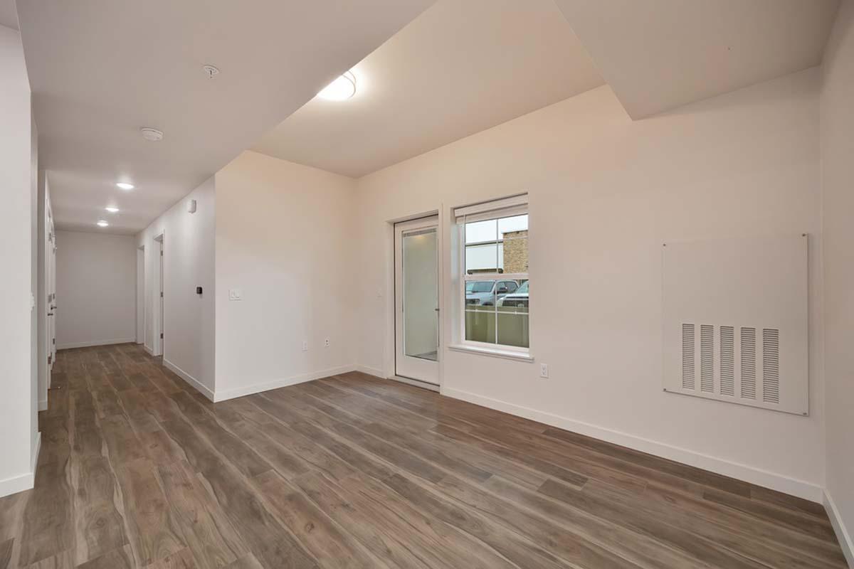 A spacious, well-lit hallway in a modern apartment featuring light wood flooring. There are several doors along the walls and a large window at the end that provides natural light. A sliding glass door leads to an outdoor area, and a wall-mounted heater is visible. The walls are painted in a neutral color.