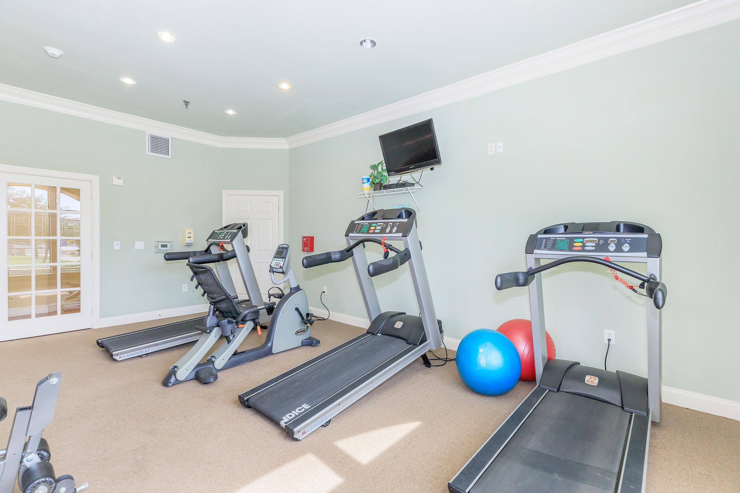 A well-lit gym area featuring two treadmills, a stationary bike, and a blue exercise ball. The walls are painted in a light green shade, and the floor is carpeted. There is a flat-screen TV mounted on the wall and a door leading to another room in the background.